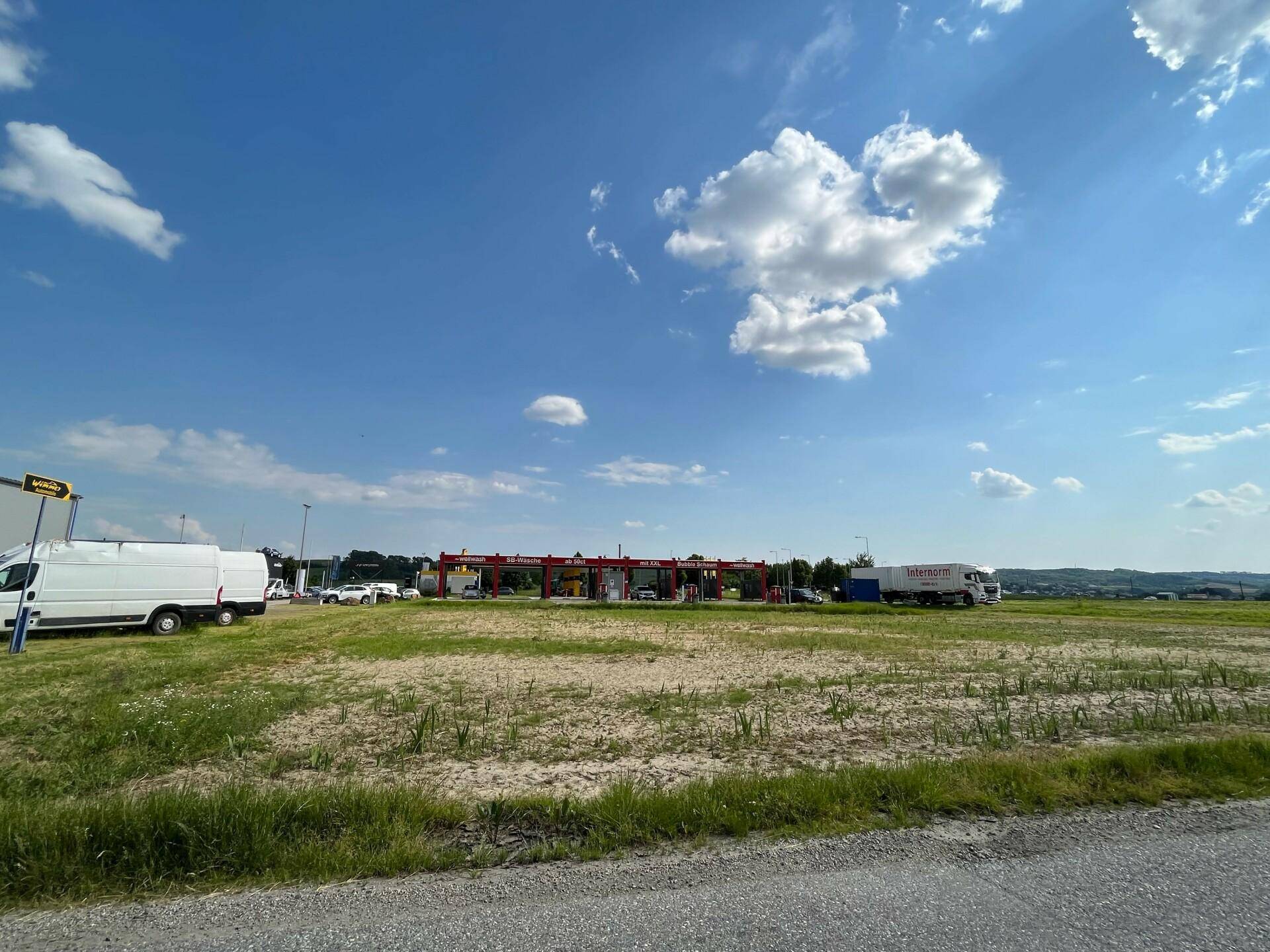 Großes, offenes Grundstück mit Gras und Schotter, im Hintergrund eine Autowaschanlage und weitere Gewerbebauten.