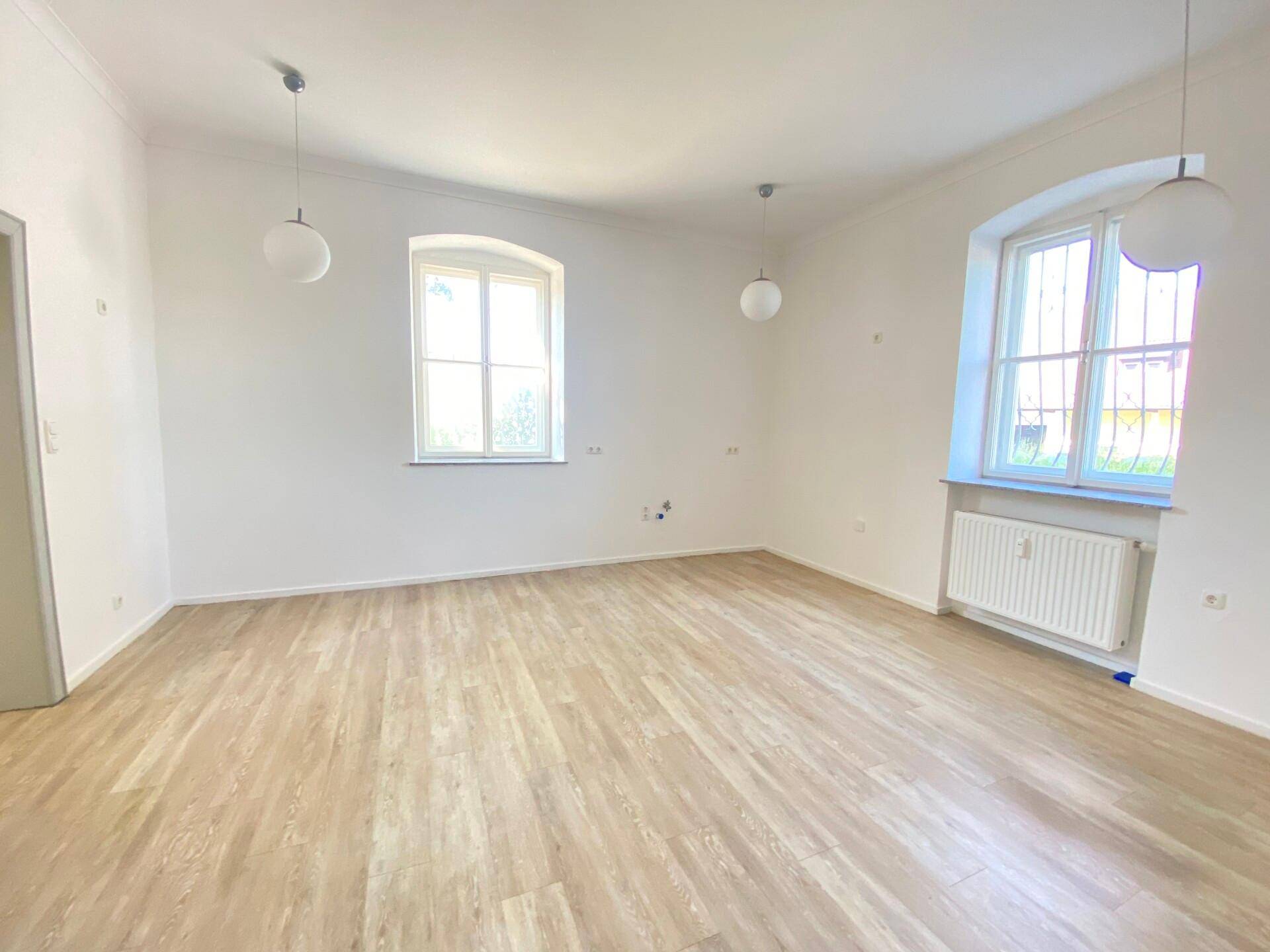 Heller, leerer Raum mit modernem Laminatboden und zwei großen Fenstern, die viel Tageslicht hereinlassen.