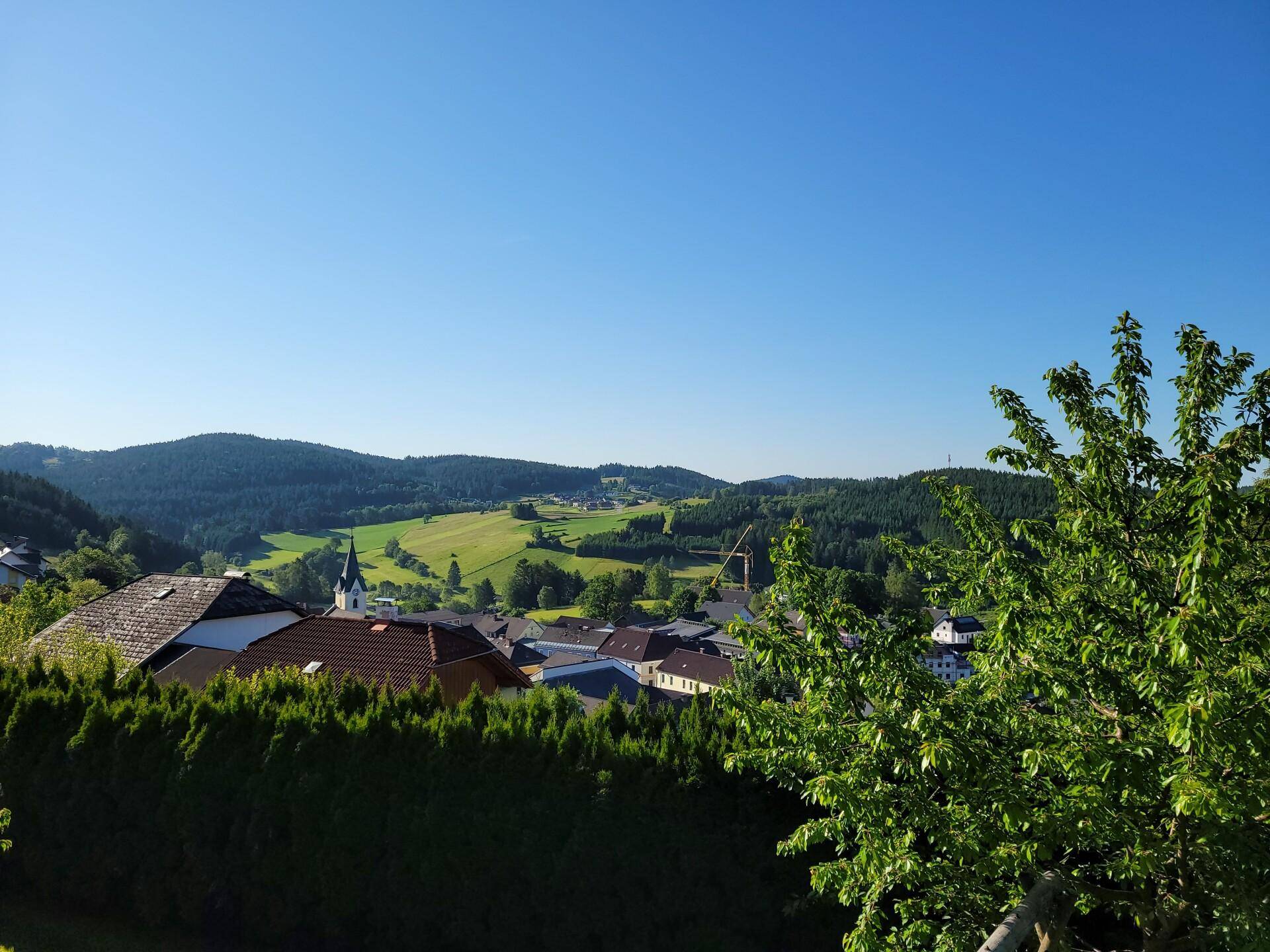 Panoramablick über eine ländliche Ortschaft mit grünen Hügeln und einem Kirchturm im Hintergrund.