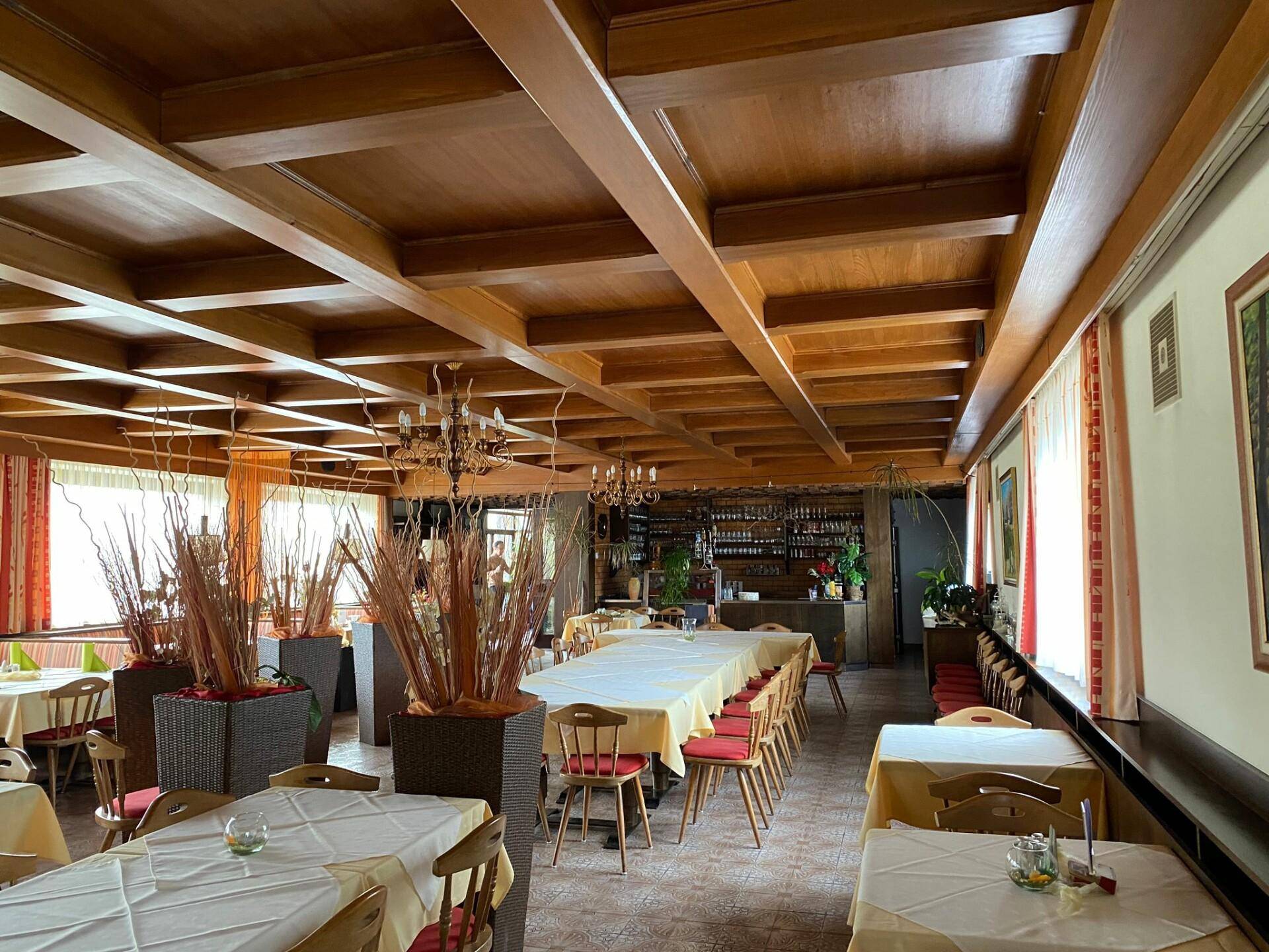 Großer Speisesaal mit Holzbalkendecke, vielen Tischen und einer Bar im Hintergrund.