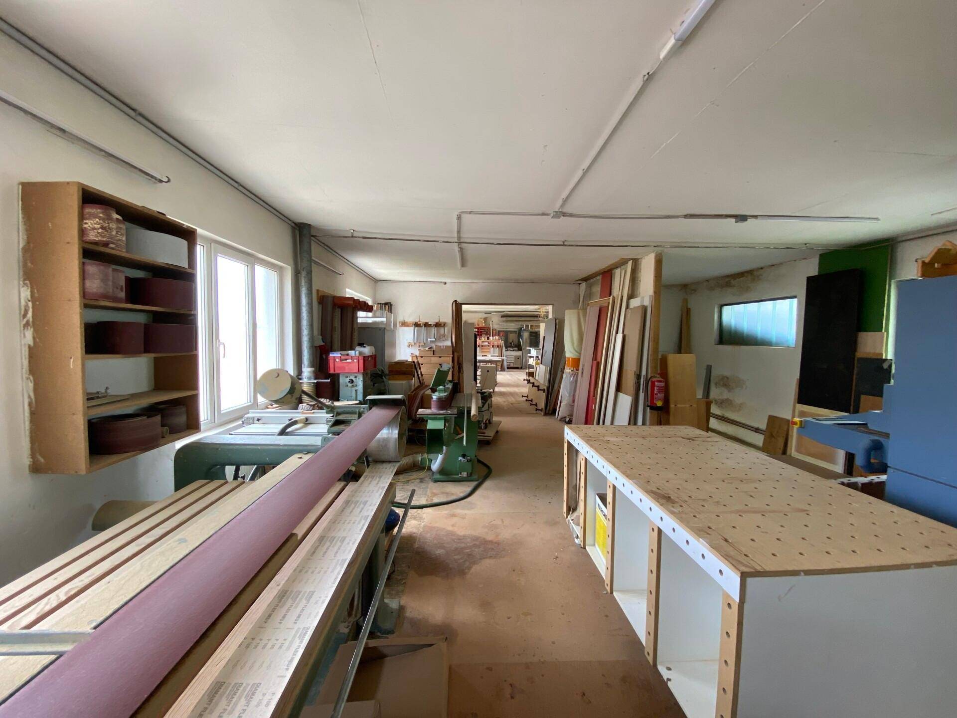 Langer Werkstattraum mit Maschinen, Arbeitsflächen und Fenstern, die viel Tageslicht hereinlassen.