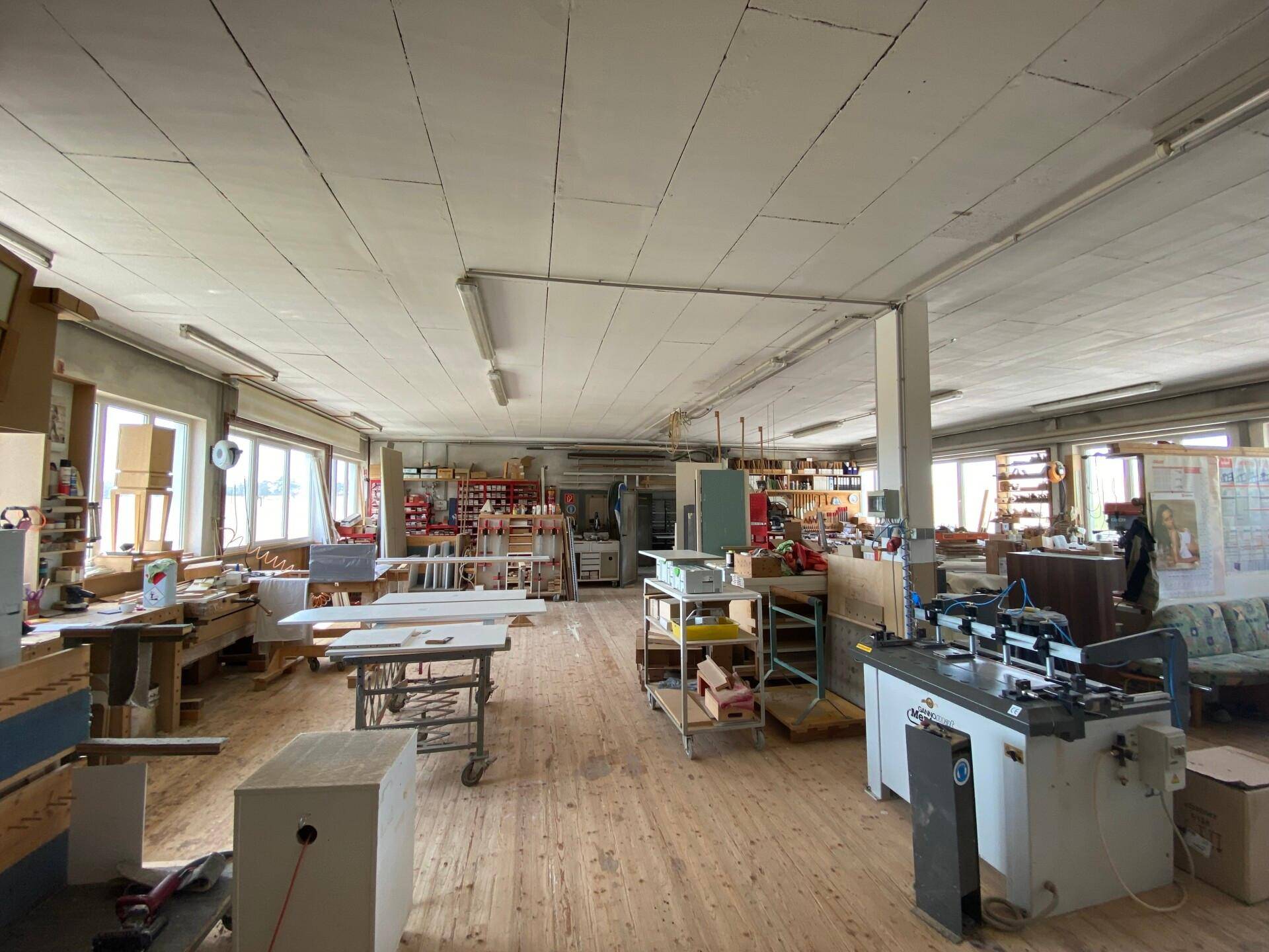 Großer, heller Werkstattraum mit Holzboden, vielen Arbeitsflächen und Maschinen unter einer hohen Decke.