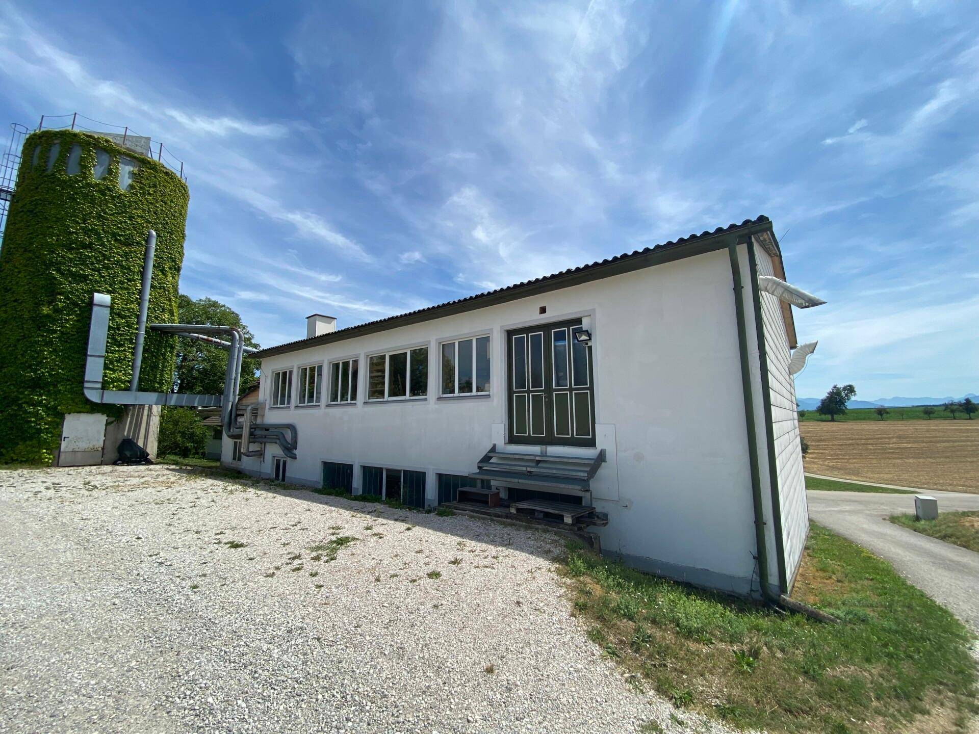 Seitenansicht eines weißen Gebäudes mit einem begrünten Silo daneben, umgeben von ländlicher Landschaft.