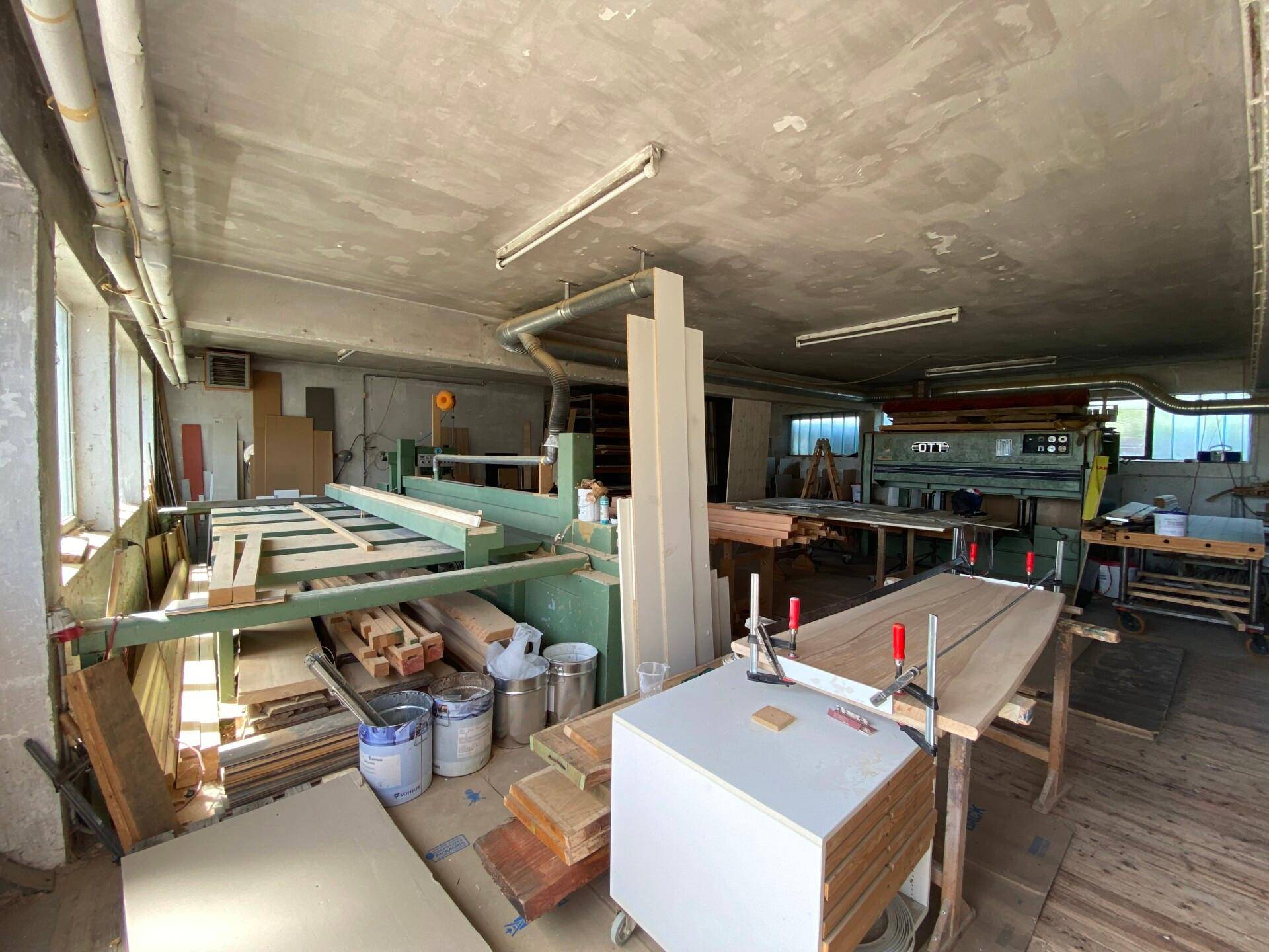 Geräumige Werkstatt mit Holzbearbeitungsmaschinen, Materiallager und hohen Decken, gut beleuchtet.