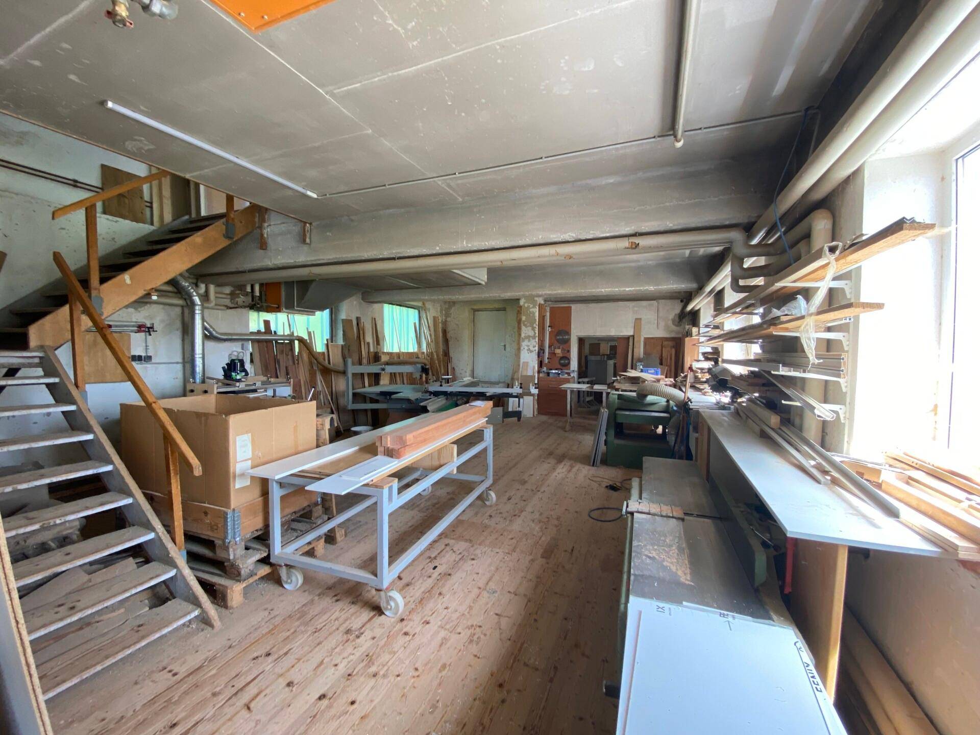 Großer Werkstattbereich mit Holztreppe, Arbeitsgeräten und gelagertem Material, mit viel Tageslicht.