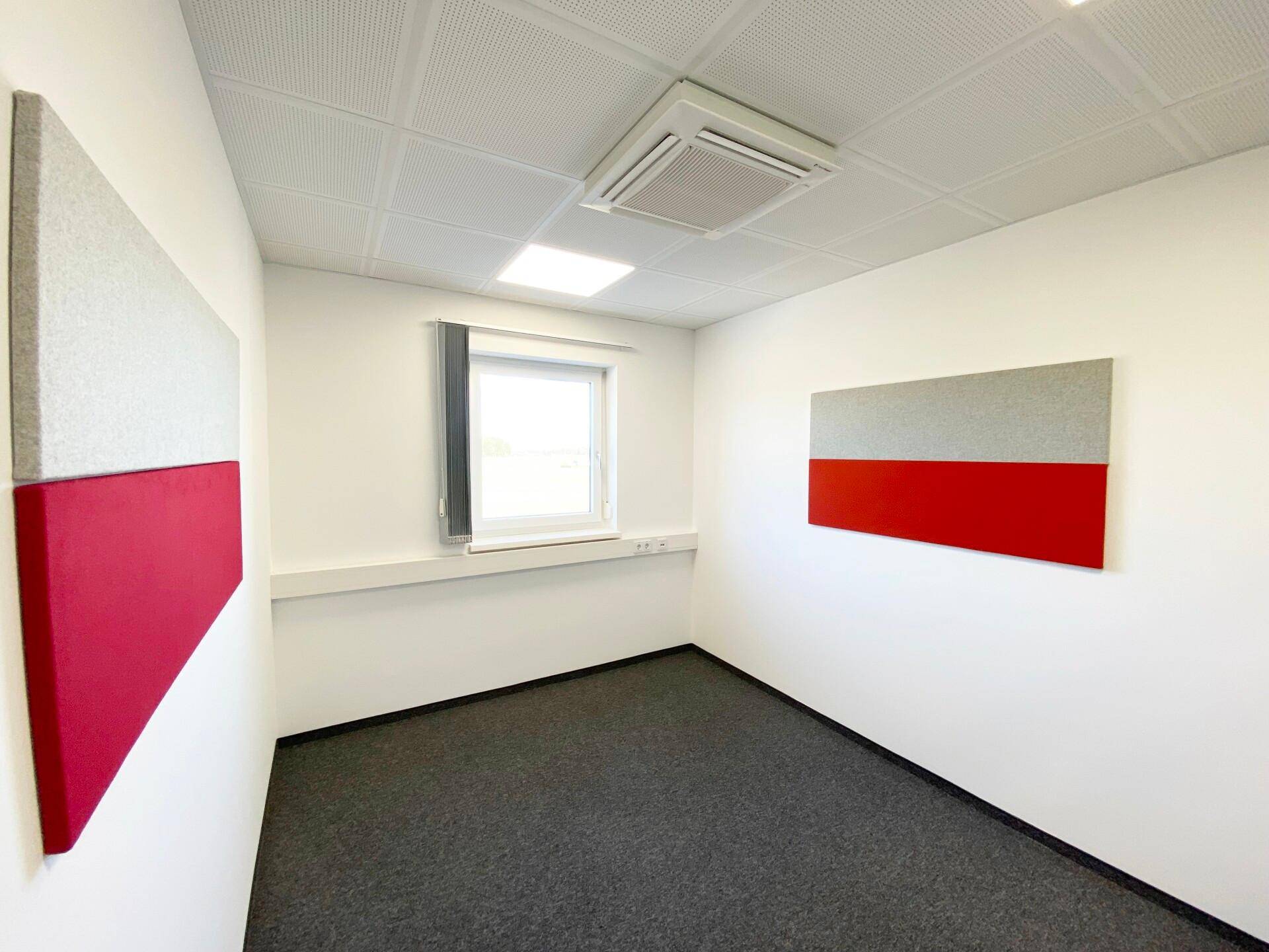 Kleineres Büro mit Teppichboden, einem Fenster und zwei rot-grauen Akustikpaneelen an den Wänden.
