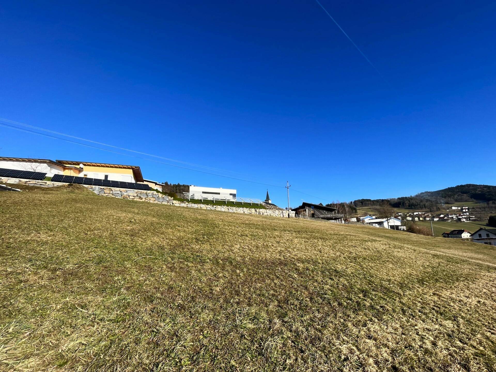 Sonnige Hanglage mit weitem Blick über grüne Wiesen und eine kleine Siedlung in der Ferne.