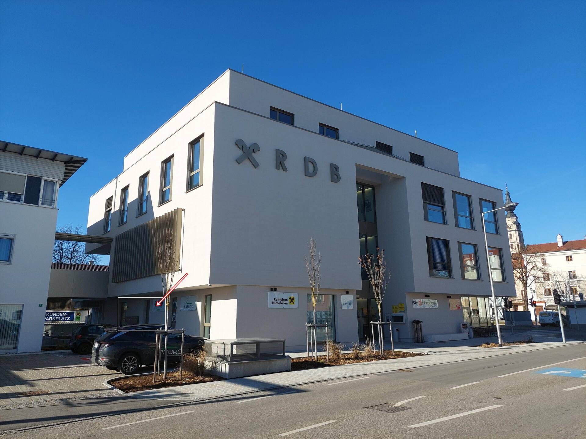 Ansicht des modernen Geschäftsgebäudes von der Straßenseite mit angrenzendem Parkplatz und Turm im Hintergrund.