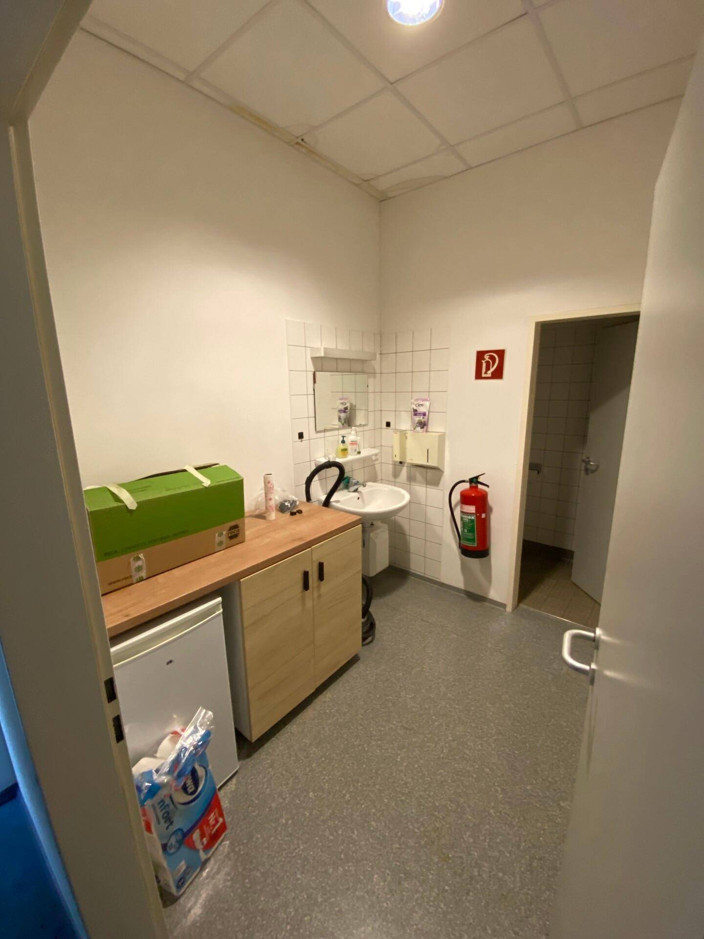Kleiner Pausenraum mit Waschbecken, Arbeitsplatte, Kühlschrank und Zugang zu weiteren Sanitärräumen.
