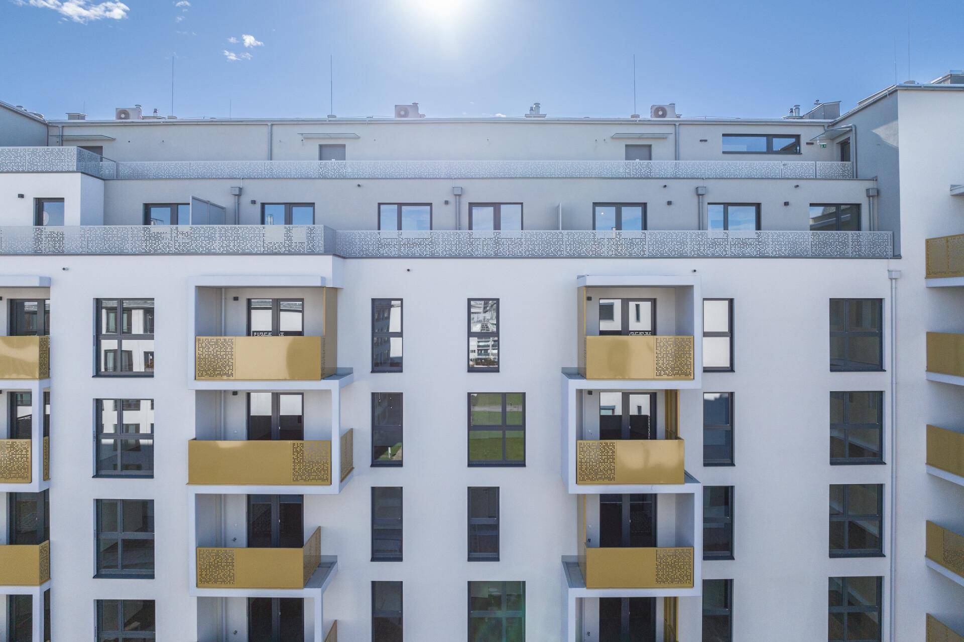 Moderne Fassade eines Wohngebäudes mit zahlreichen Balkonen und dekorativen goldenen Geländern unter blauem Himmel.