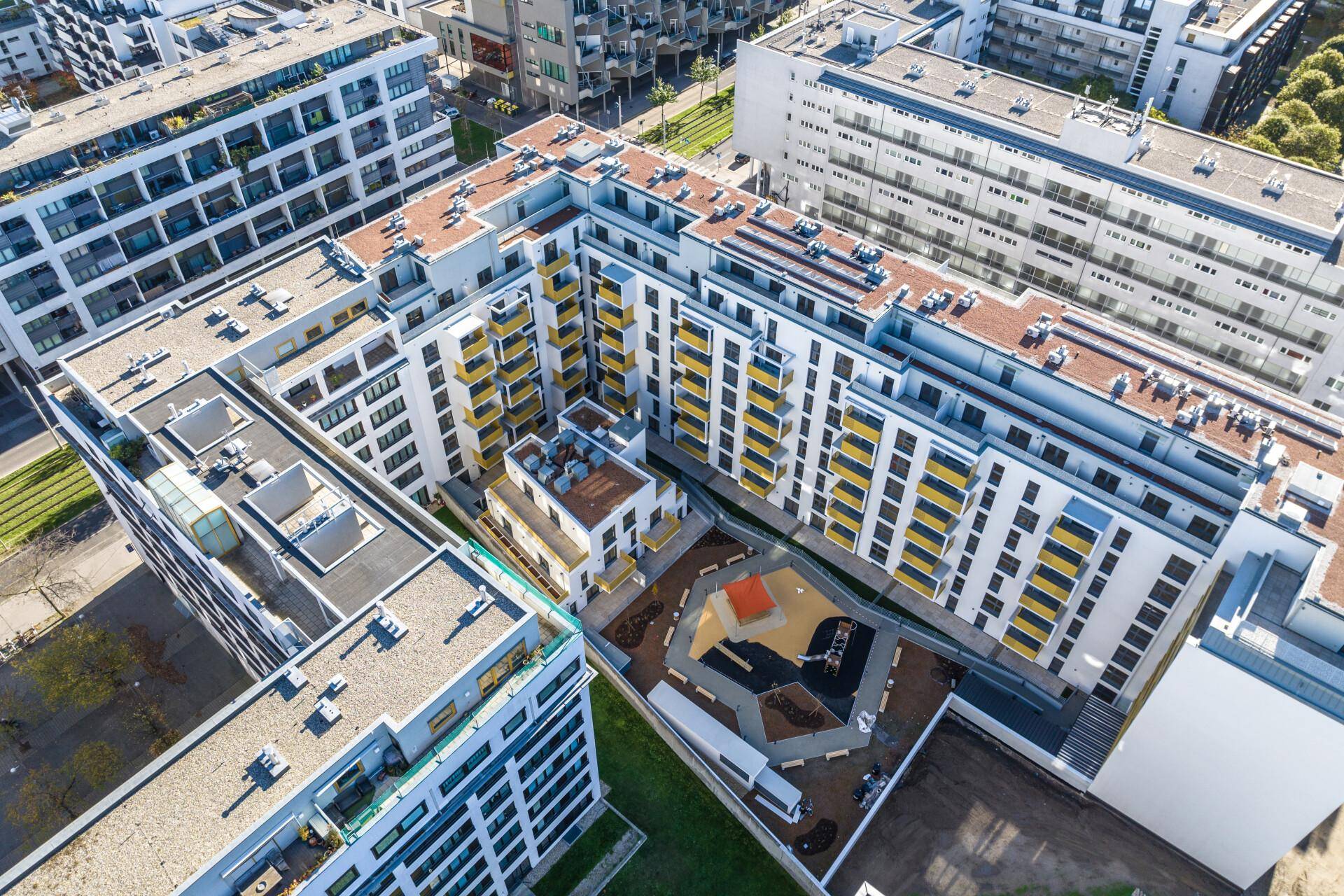 Luftaufnahme eines modernen Wohnkomplexes mit Innenhof, Spielplatz und umliegenden Grünflächen.