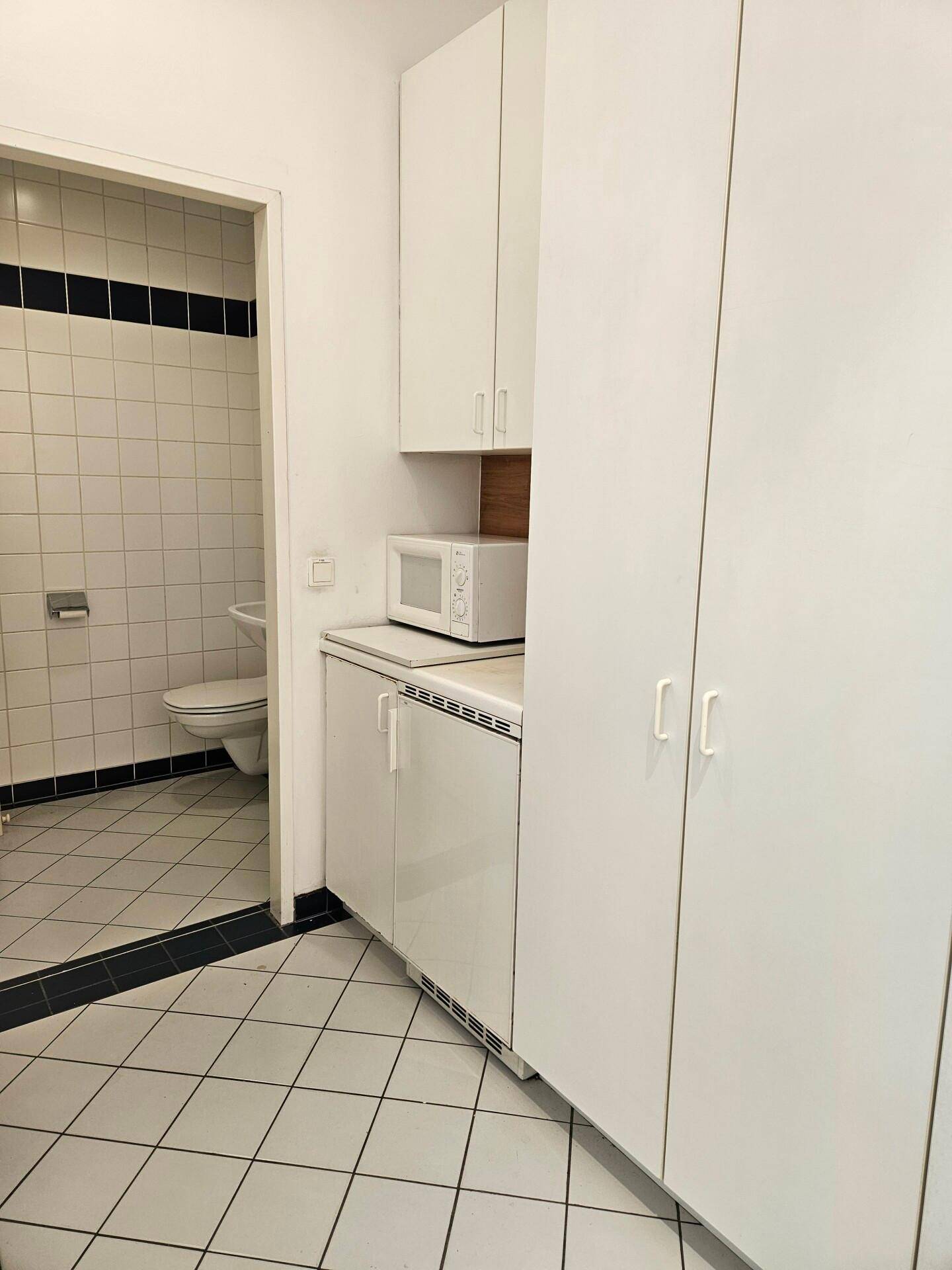 Kleine Küchenzeile mit weißen Schränken, Mikrowelle und Zugang zu einem gefliesten Badezimmer.