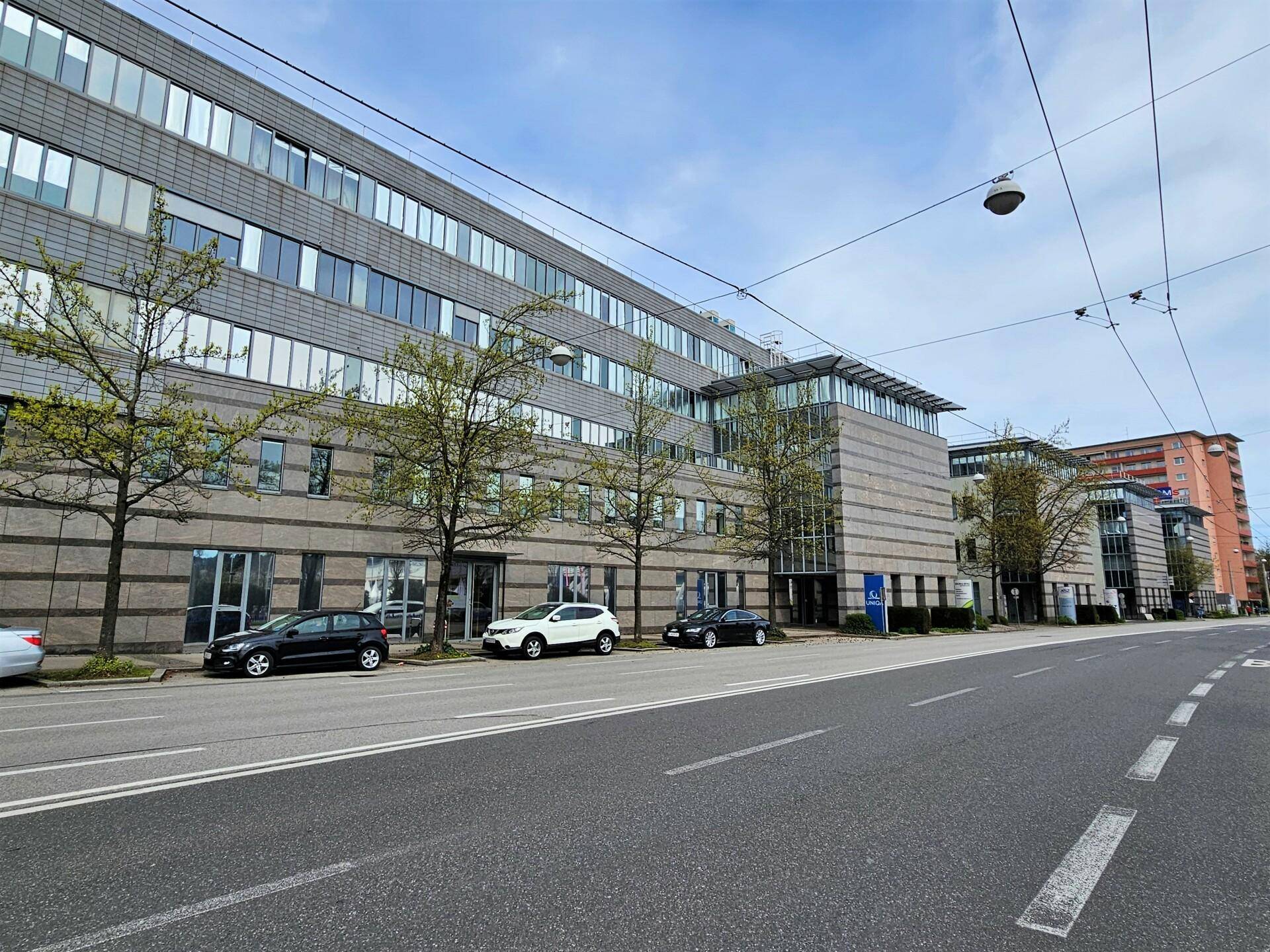 Moderne Bürogebäude entlang einer Straße mit parkenden Autos und Bäumen unter blauem Himmel.