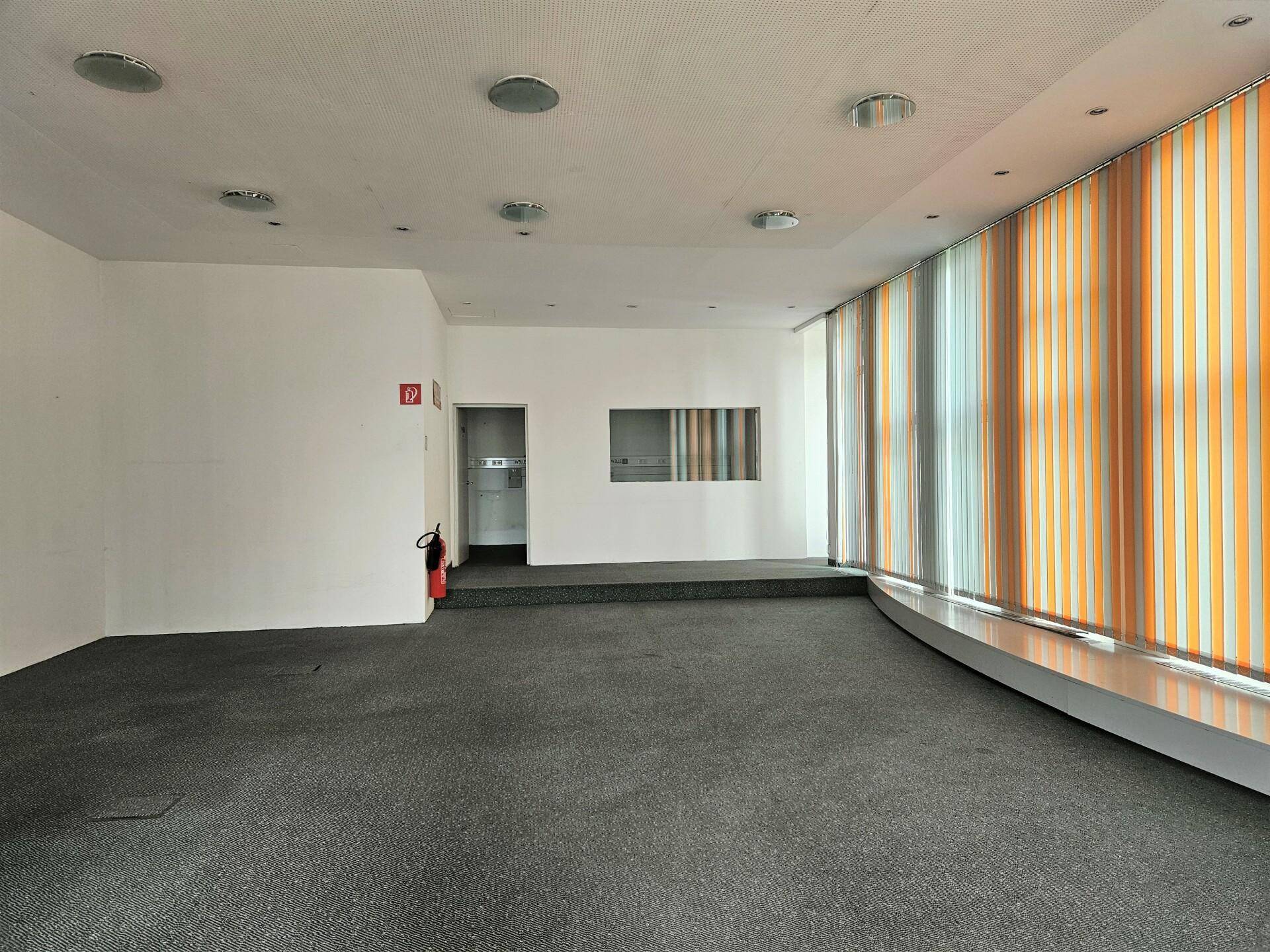 Heller Büroraum mit Teppichboden, einer erhöhten Plattform und vertikalen Jalousien an den Fenstern.