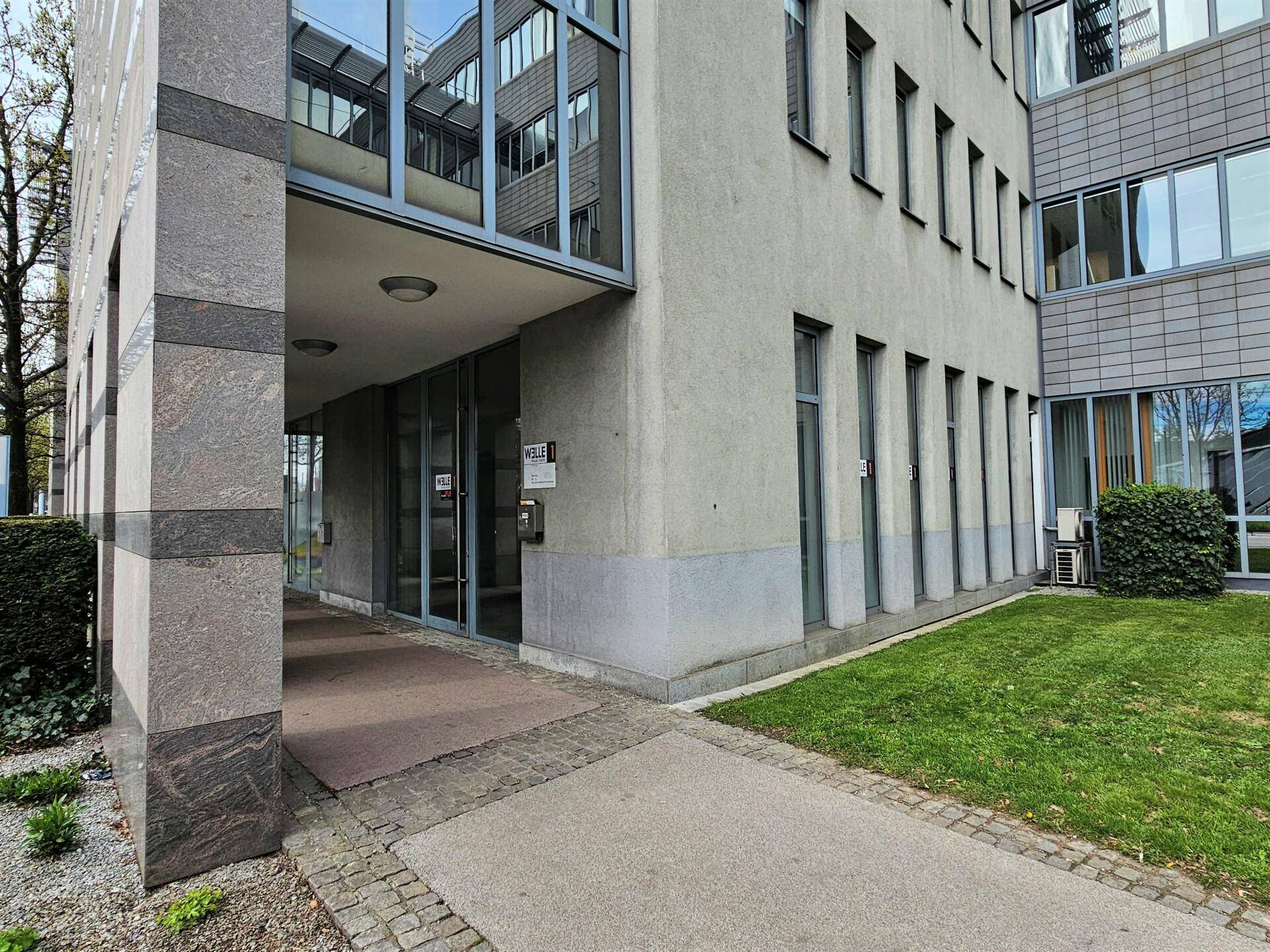Eingangsbereich eines modernen Bürogebäudes mit Glasfassade und gepflegter Grünfläche.