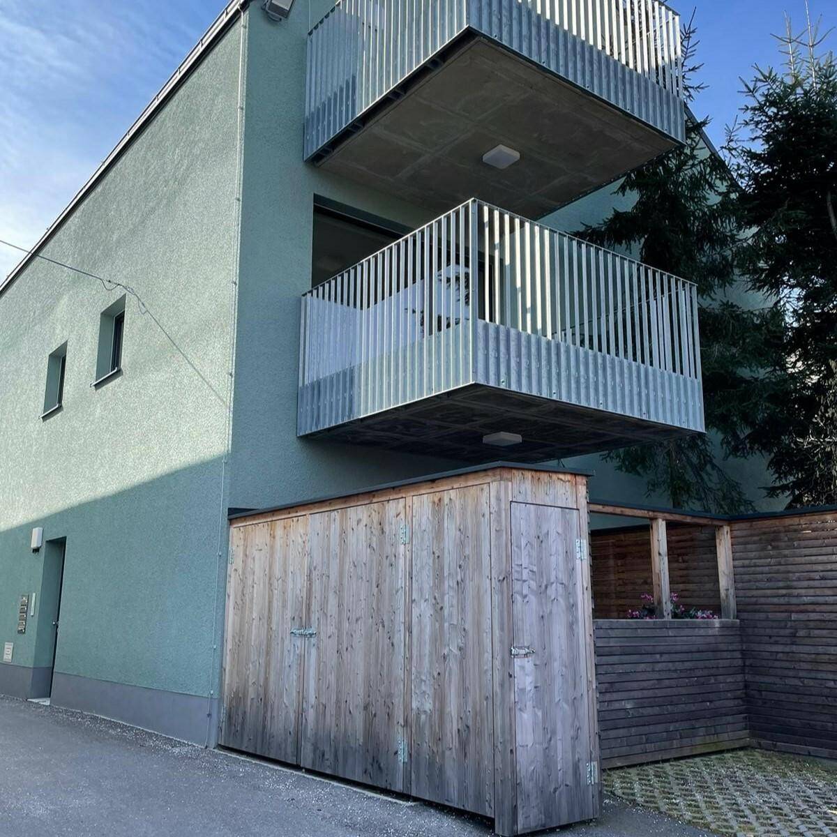 Moderne Gebäudefassade in Grün mit mehreren Balkonen und Holzverkleidung im Erdgeschoss.