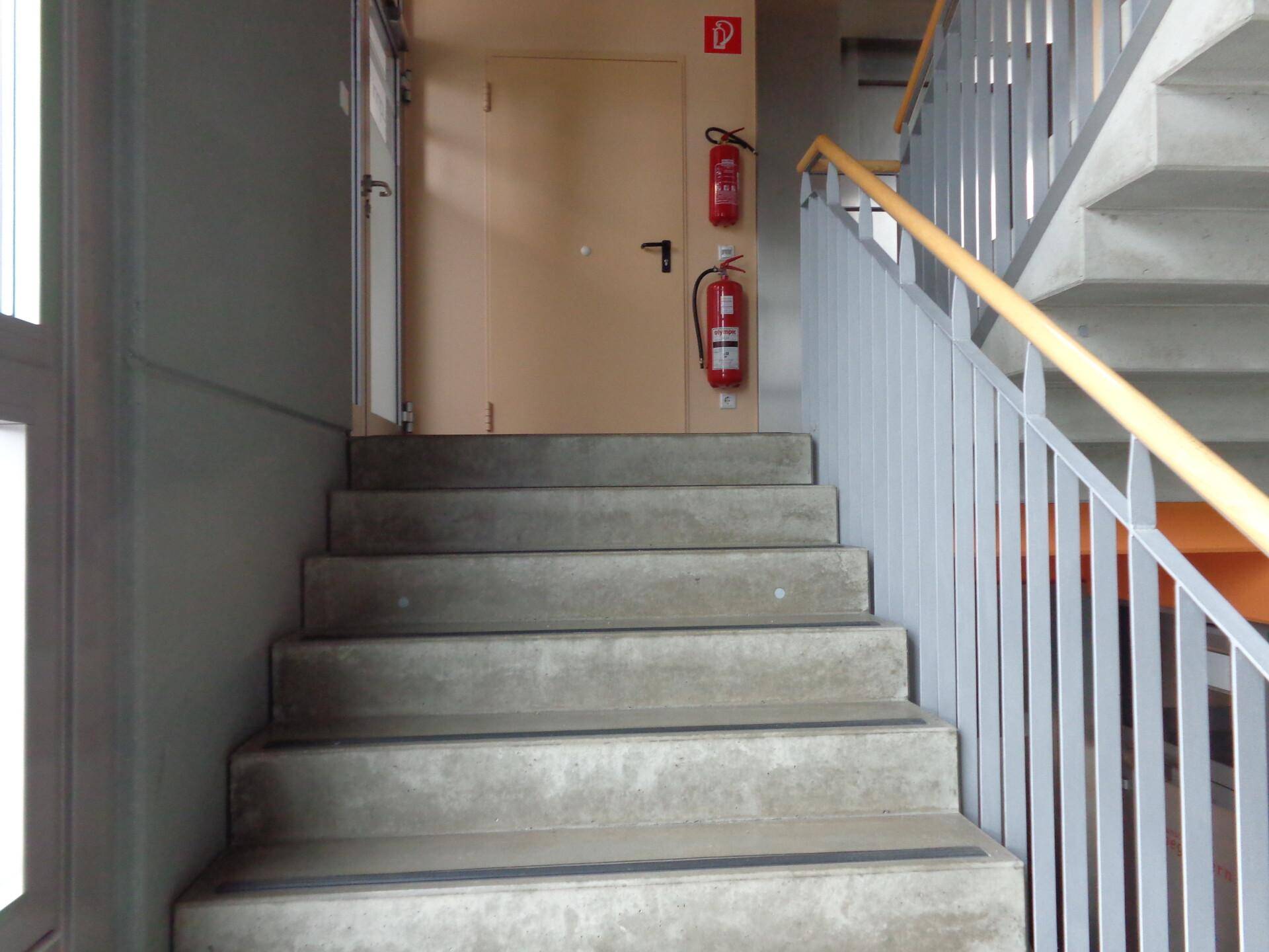 Robuste Betontreppe mit Metallgeländer und Holzhandlauf, ausgestattet mit Feuerlöschern.