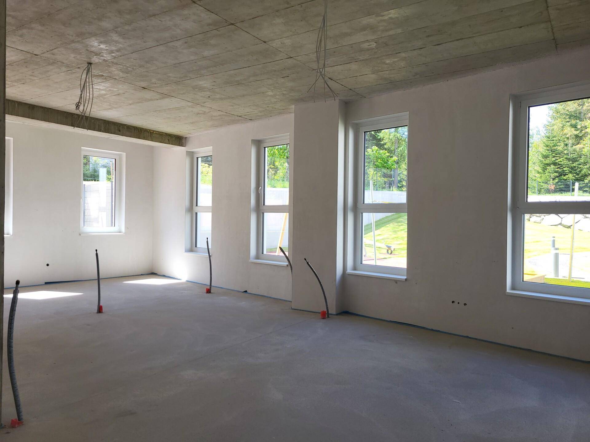 Unfertiger Innenraum mit Betonboden und -decke, weißen Wänden und großen Fenstern mit Blick ins Grüne.
