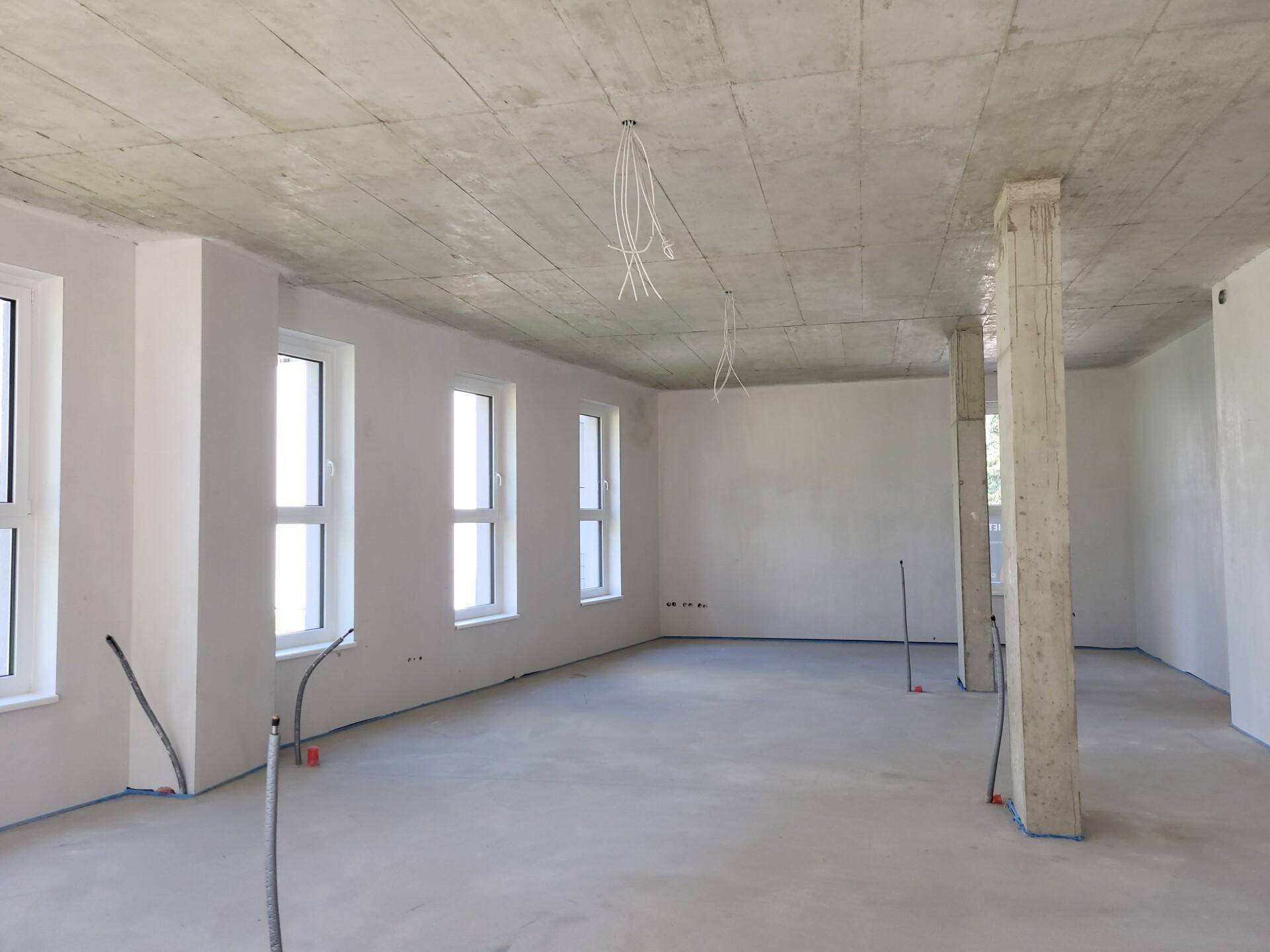 Großer, unfertiger Raum mit Betonstütze, hohen Decken und vielen Fenstern, die viel Tageslicht hereinlassen.