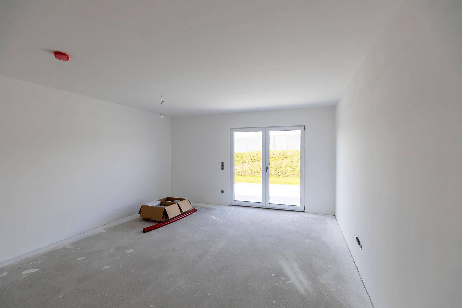 Helles, unfertiges Zimmer mit weißen Wänden, Betonboden und großer Terrassentür zum Garten.