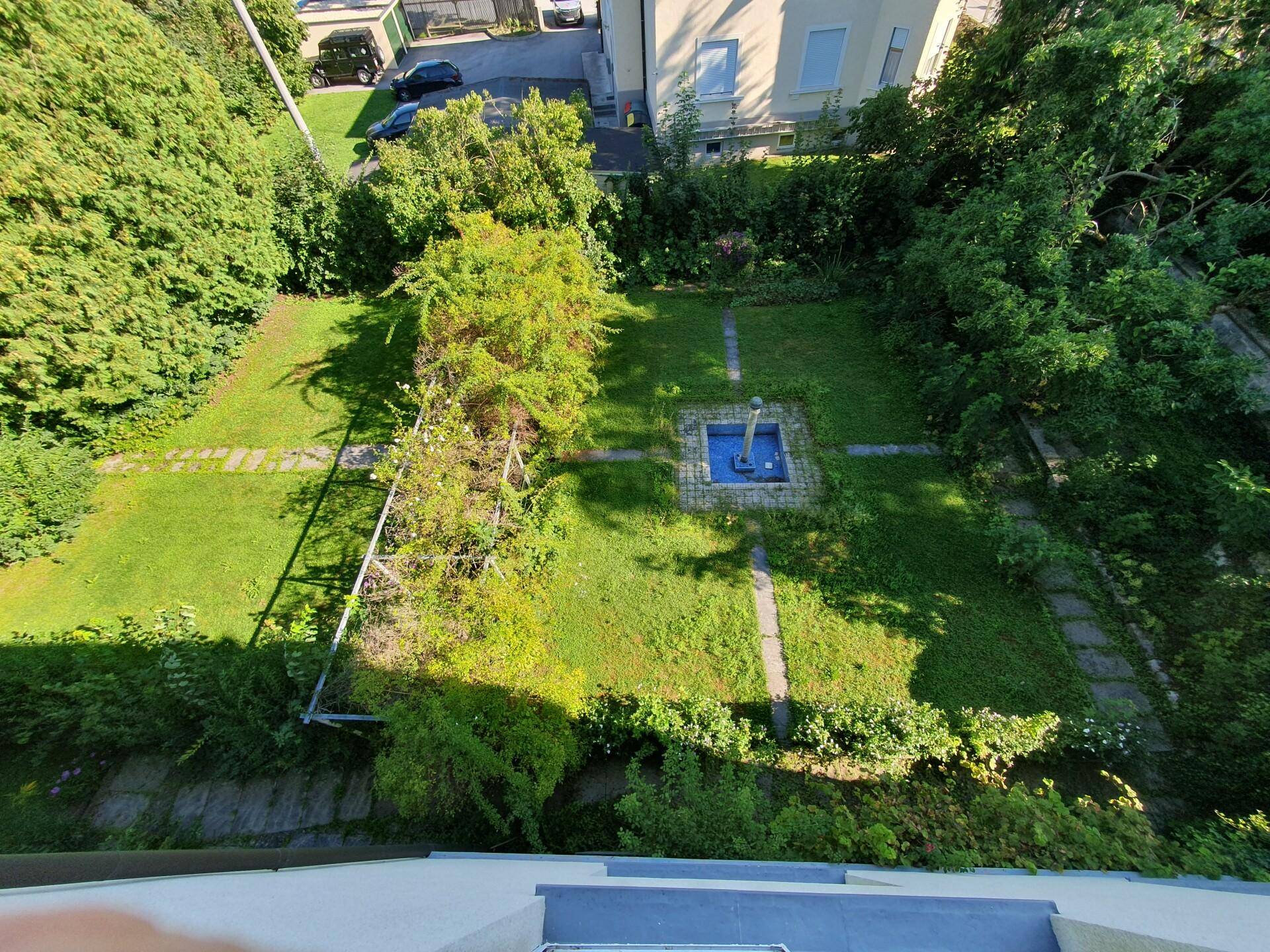 Gepflegter Gartenbereich mit Rasenflächen, Wegen und einem zentralen Brunnen, umgeben von üppigem Grün.