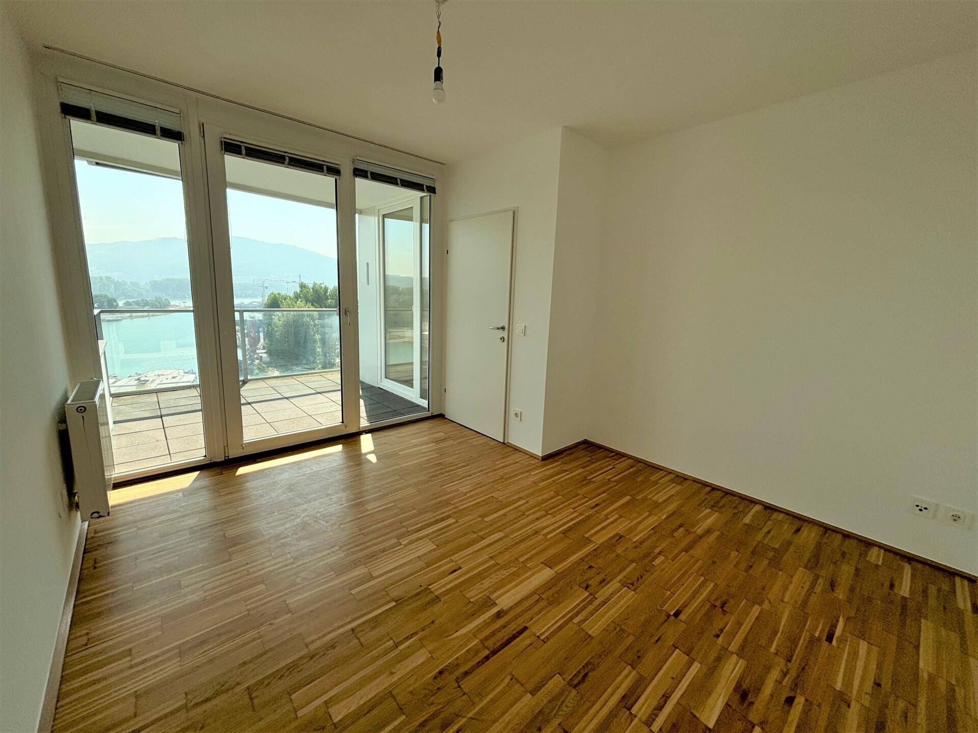 Helles Zimmer mit Holzboden und großen Glasfronten, die Zugang zu einem Außenbereich bieten.