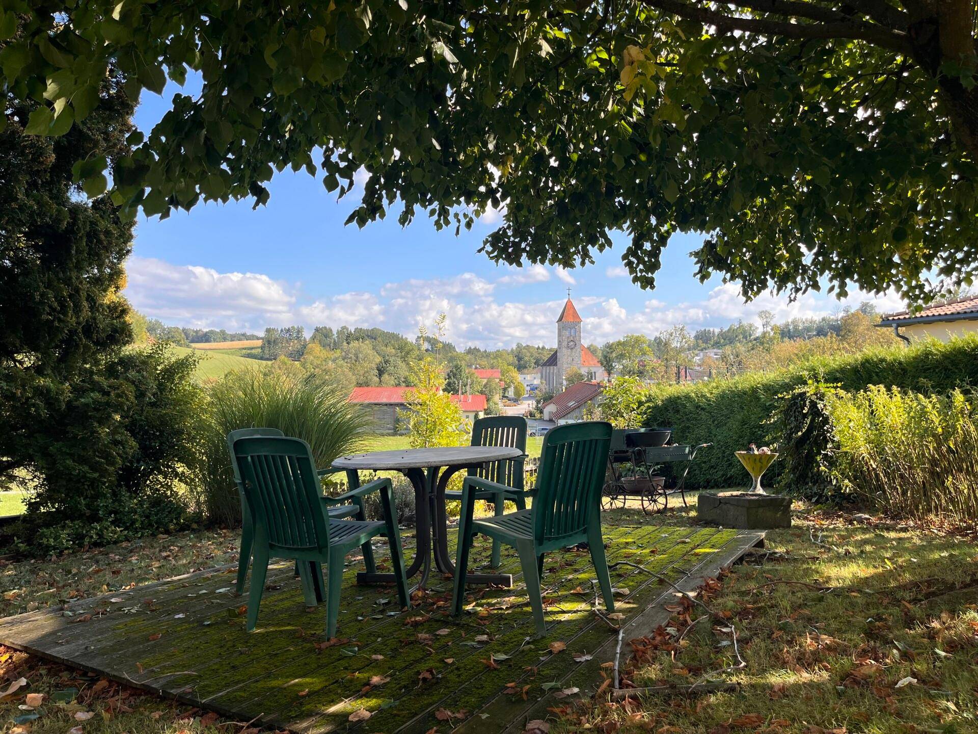 Gemütlicher Sitzbereich im Garten unter Bäumen mit Blick auf eine Kirche und die Landschaft.