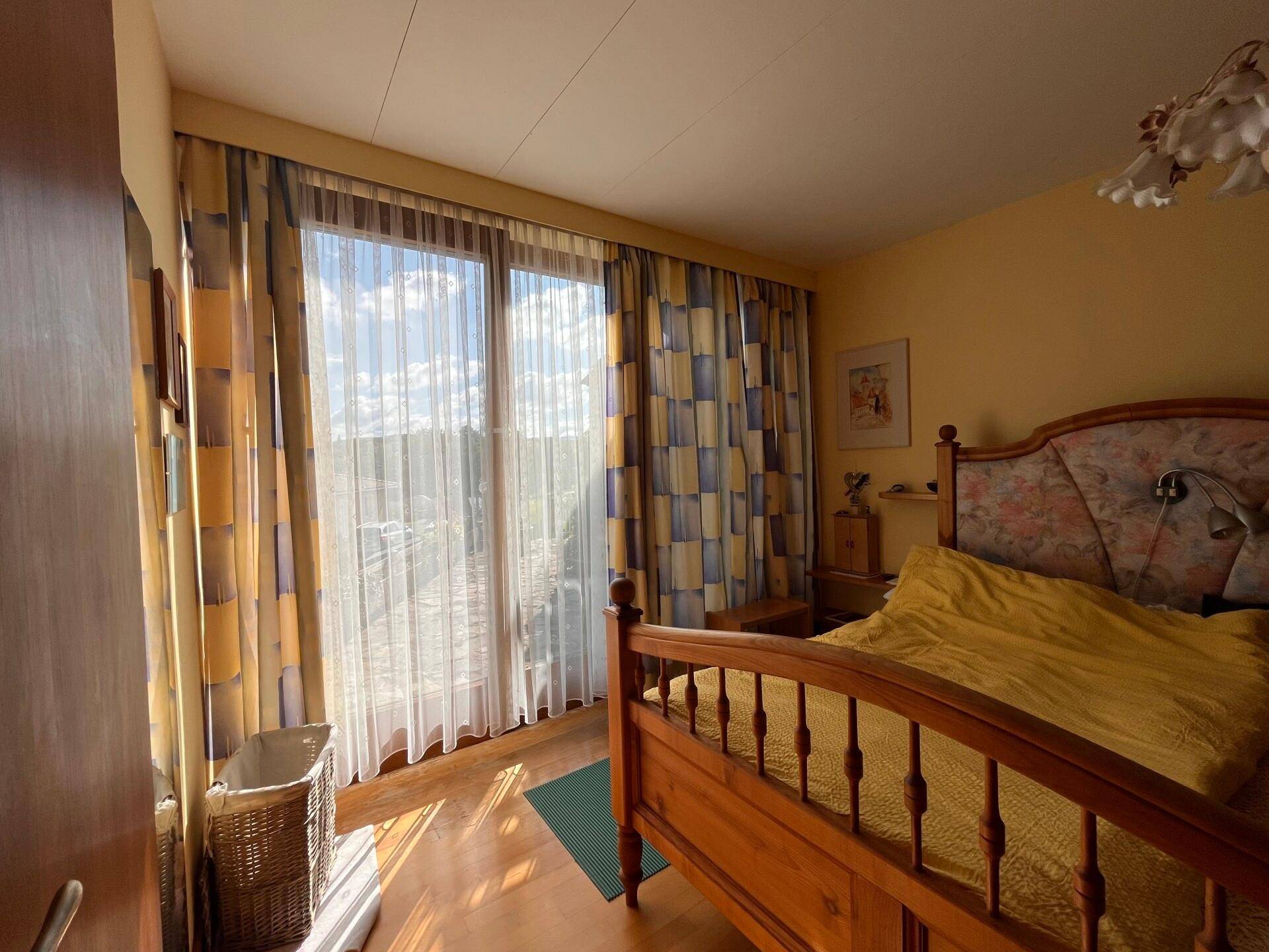 Helles Schlafzimmer mit Holzbett, Parkettboden und großen Fenstern mit Vorhängen.
