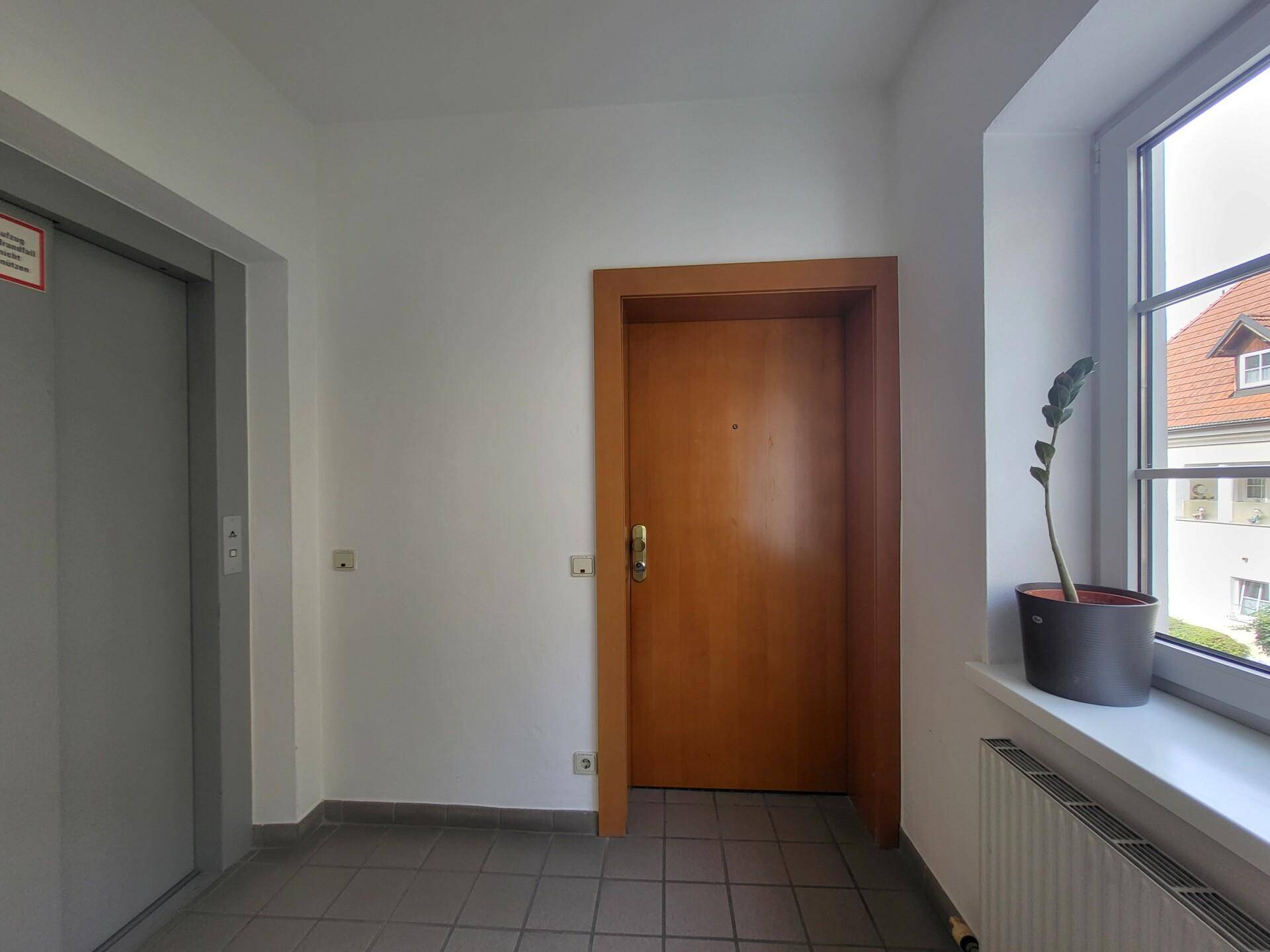 Heller Flur mit Fliesenboden, Aufzug und einer Holztür, die zu einer Wohneinheit führt.