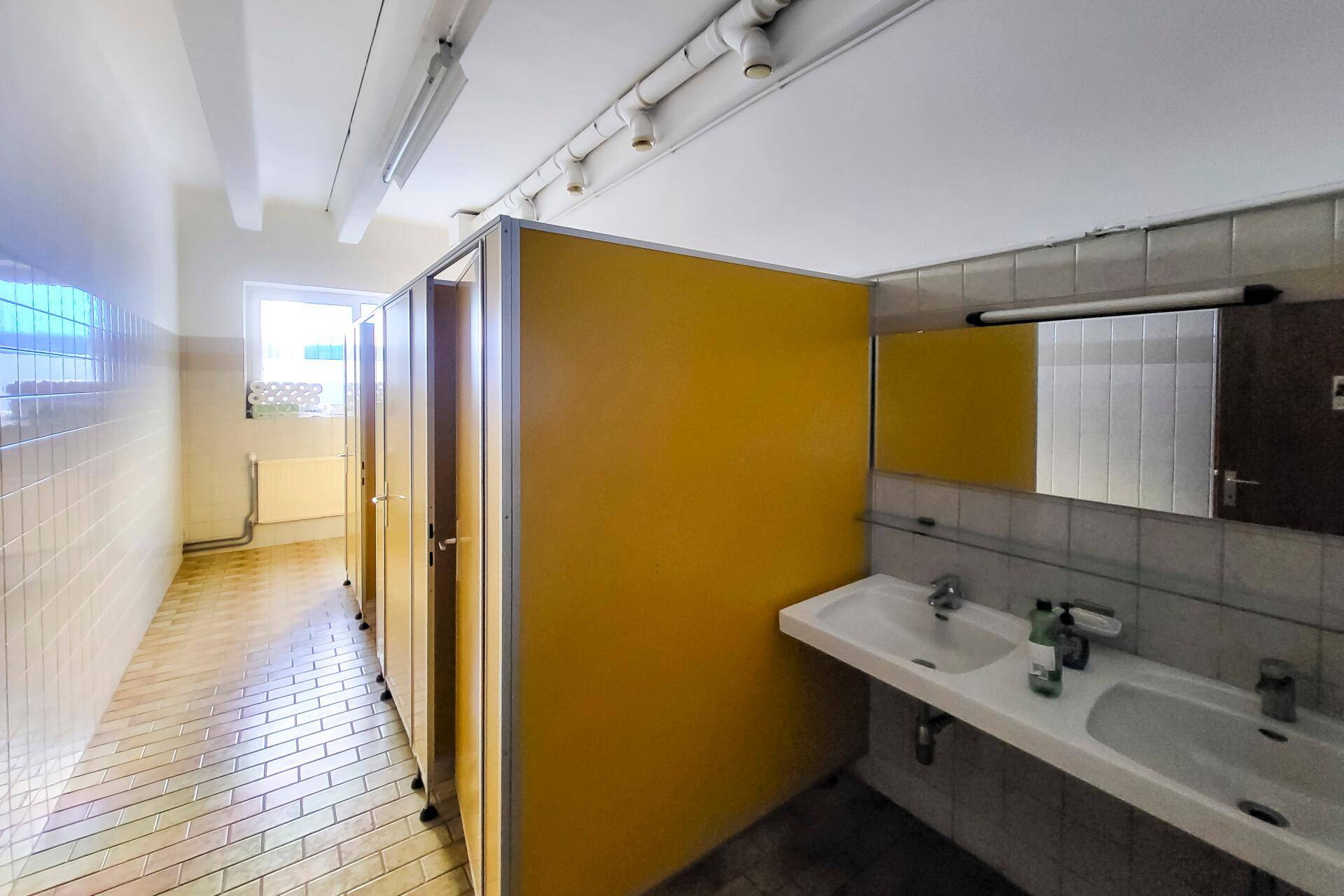 Sanitärbereich mit mehreren Toilettenkabinen, Doppelwaschbecken und einem Fenster für natürliche Belüftung.