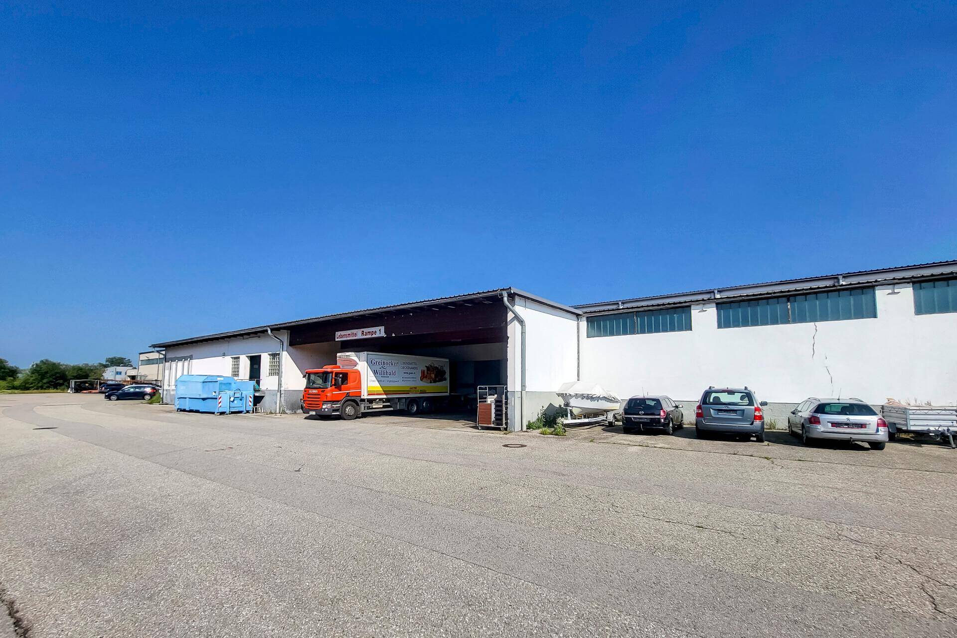 Außenansicht einer großen Lagerhalle mit Laderampen und Parkplätzen unter blauem Himmel.