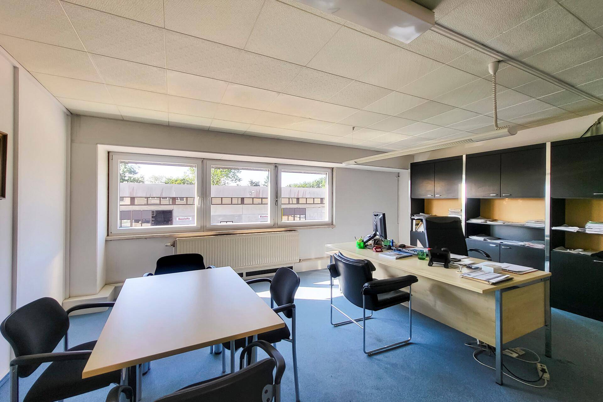 Geräumiges Büro mit Teppichboden, großen Fenstern und ausreichend Platz für Besprechungen und Arbeitsplätze.