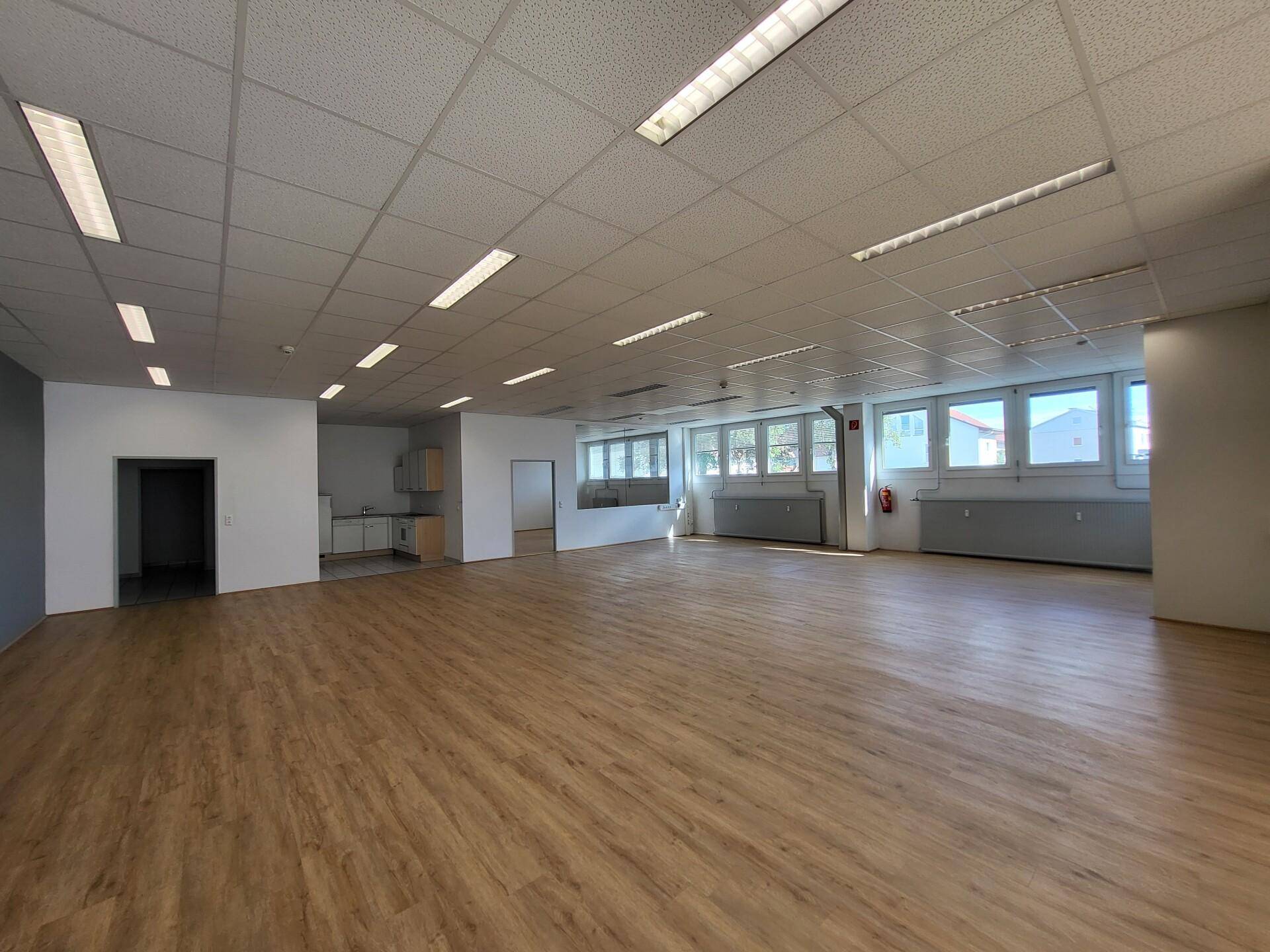 Großer, offener Gewerberaum mit hellem Laminatboden, abgehängter Decke und einer kleinen Teeküche im Hintergrund.