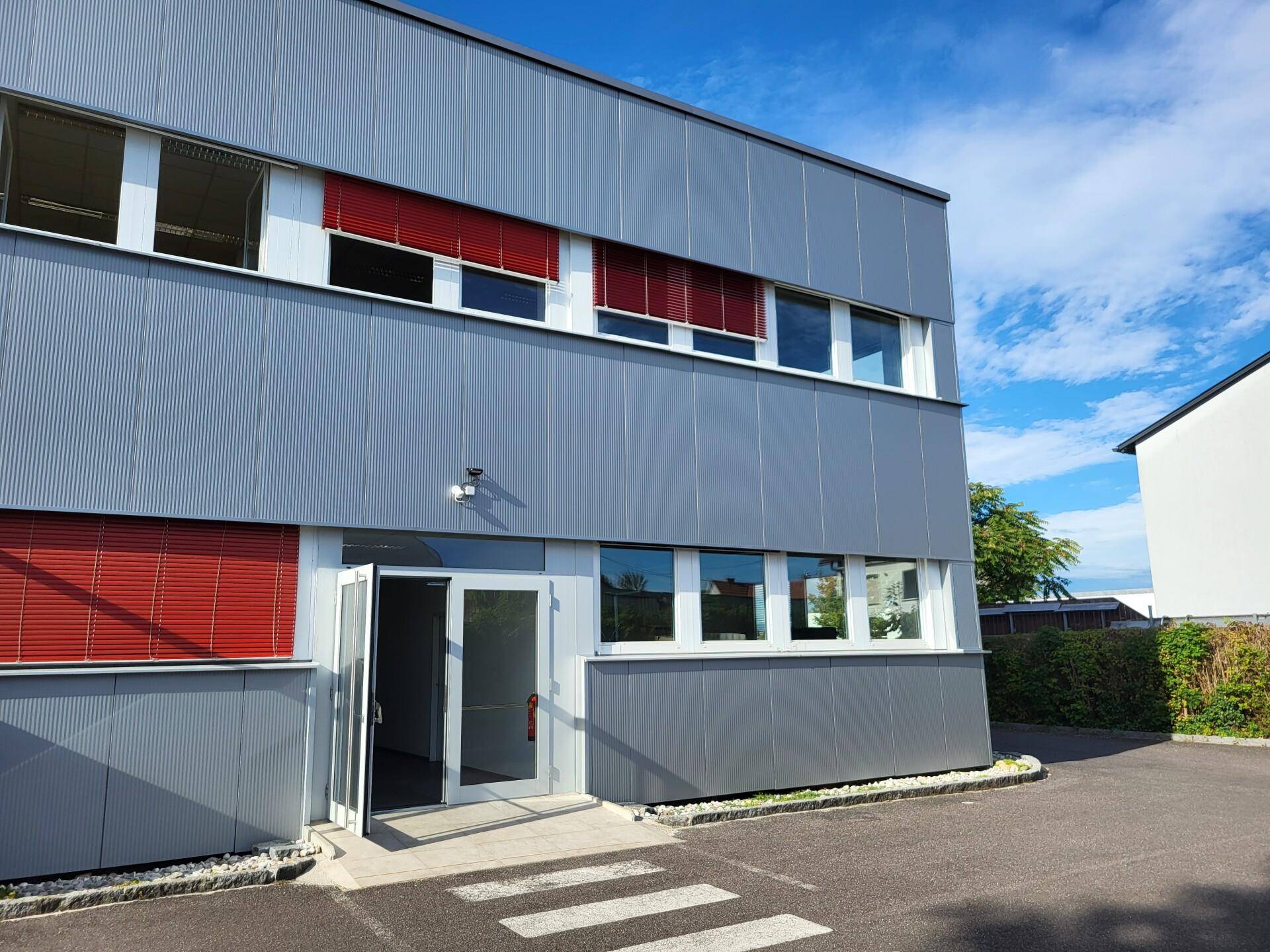 Modernes Gebäude mit grauer Wellblechfassade, roten Jalousien und gläsernem Eingangsbereich unter blauem Himmel.