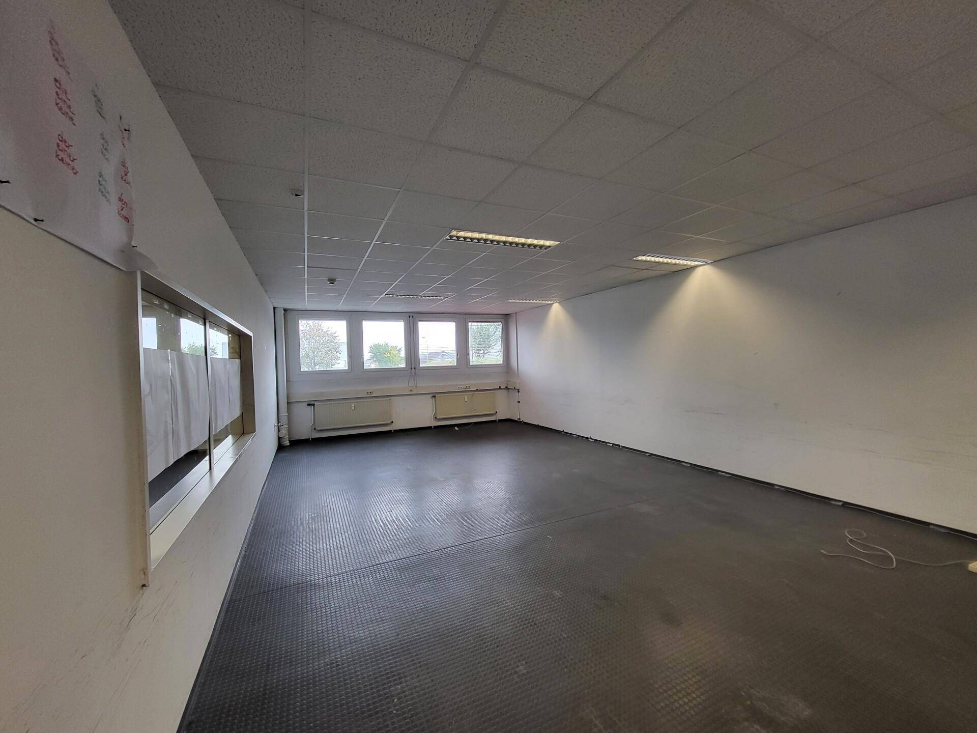 Langer Gewerberaum mit dunklem Vinylboden, weißen Wänden und einer Fensterfront, die viel Licht hereinlässt.