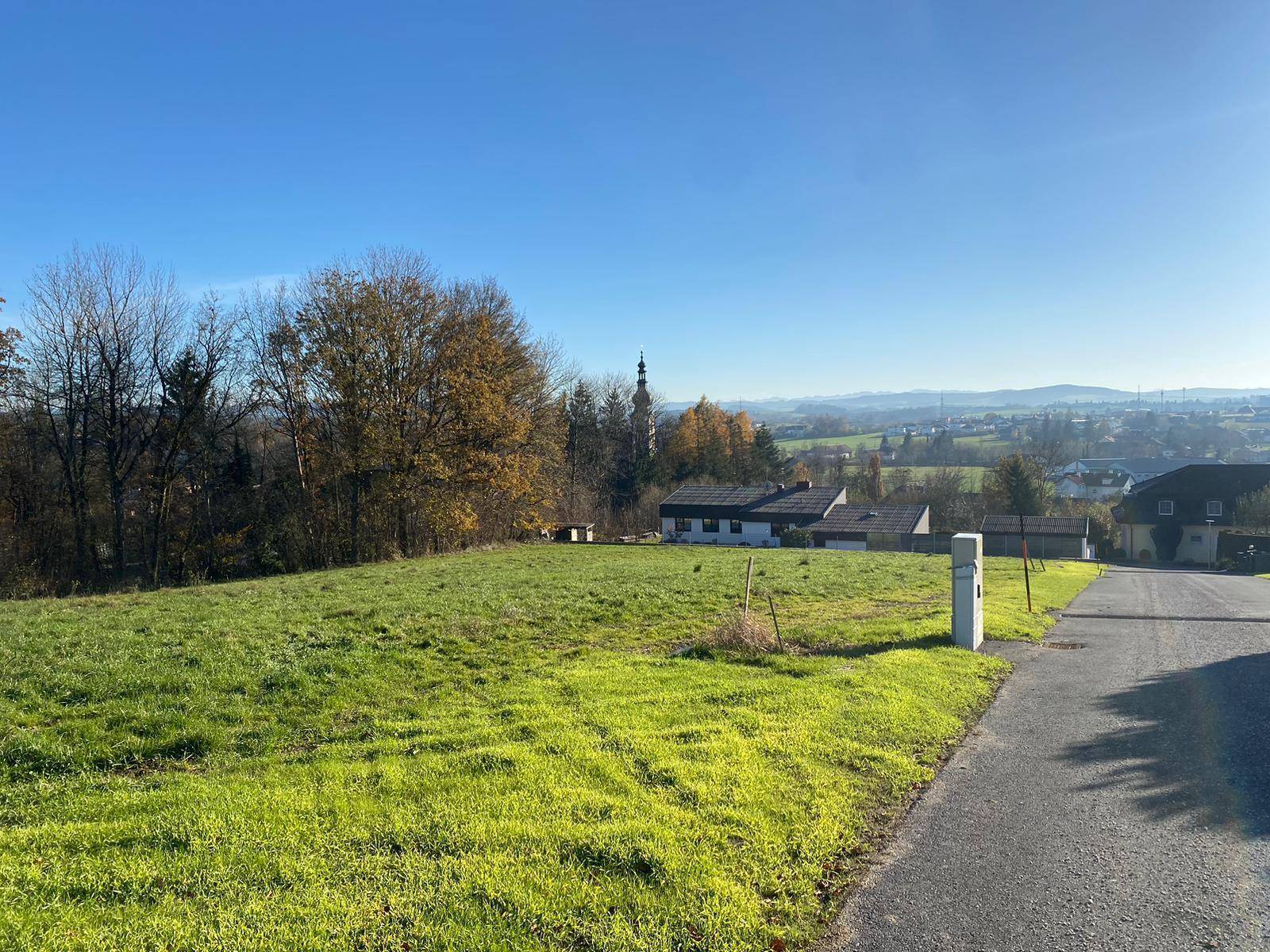 Sonnige Grünfläche mit Zufahrtsweg und weitem Blick über die Landschaft und angrenzende Bebauung.