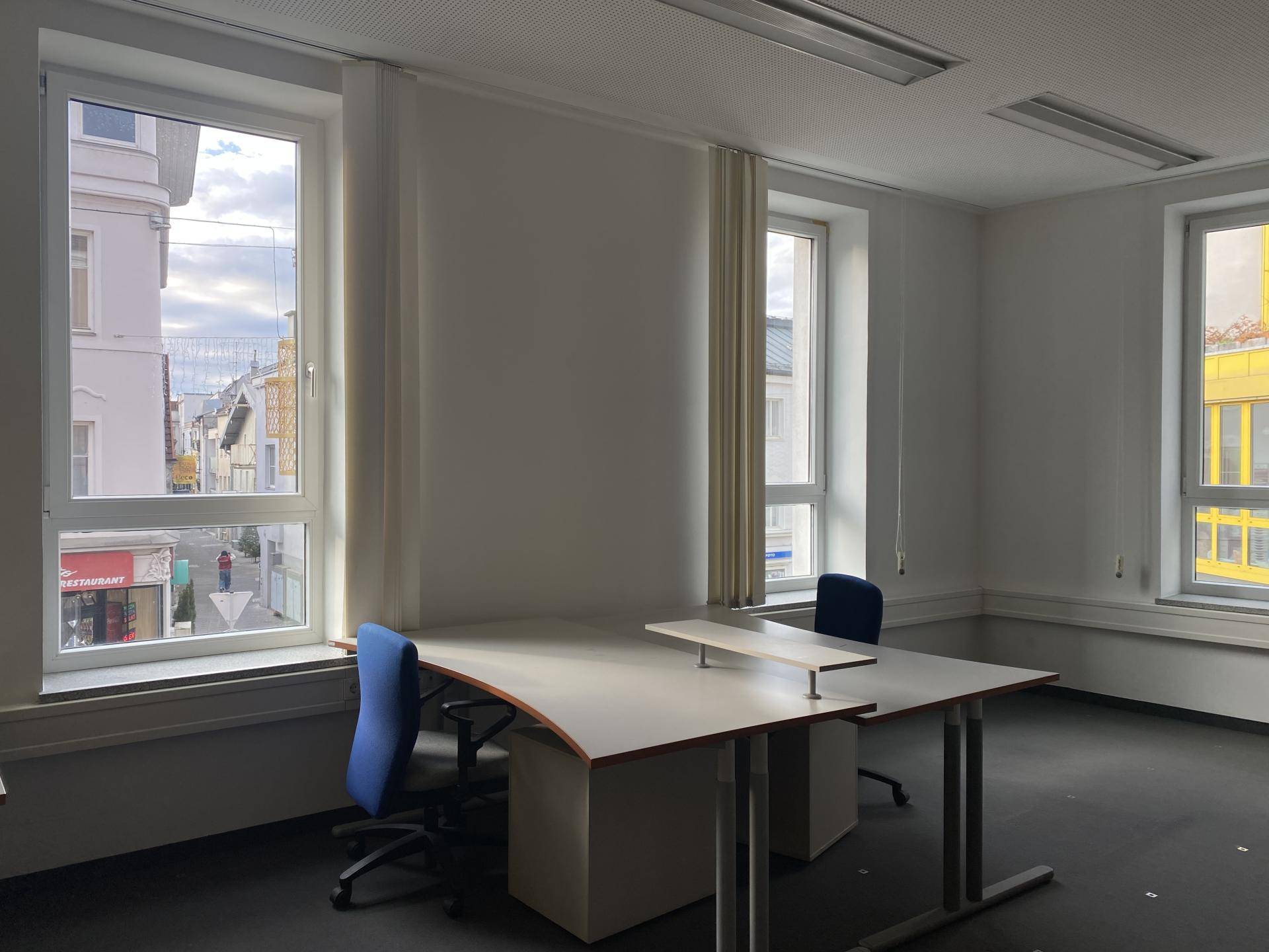Heller Büroraum mit mehreren Arbeitsplätzen und Blick auf die Stadt durch große Fenster, gut beleuchtet.