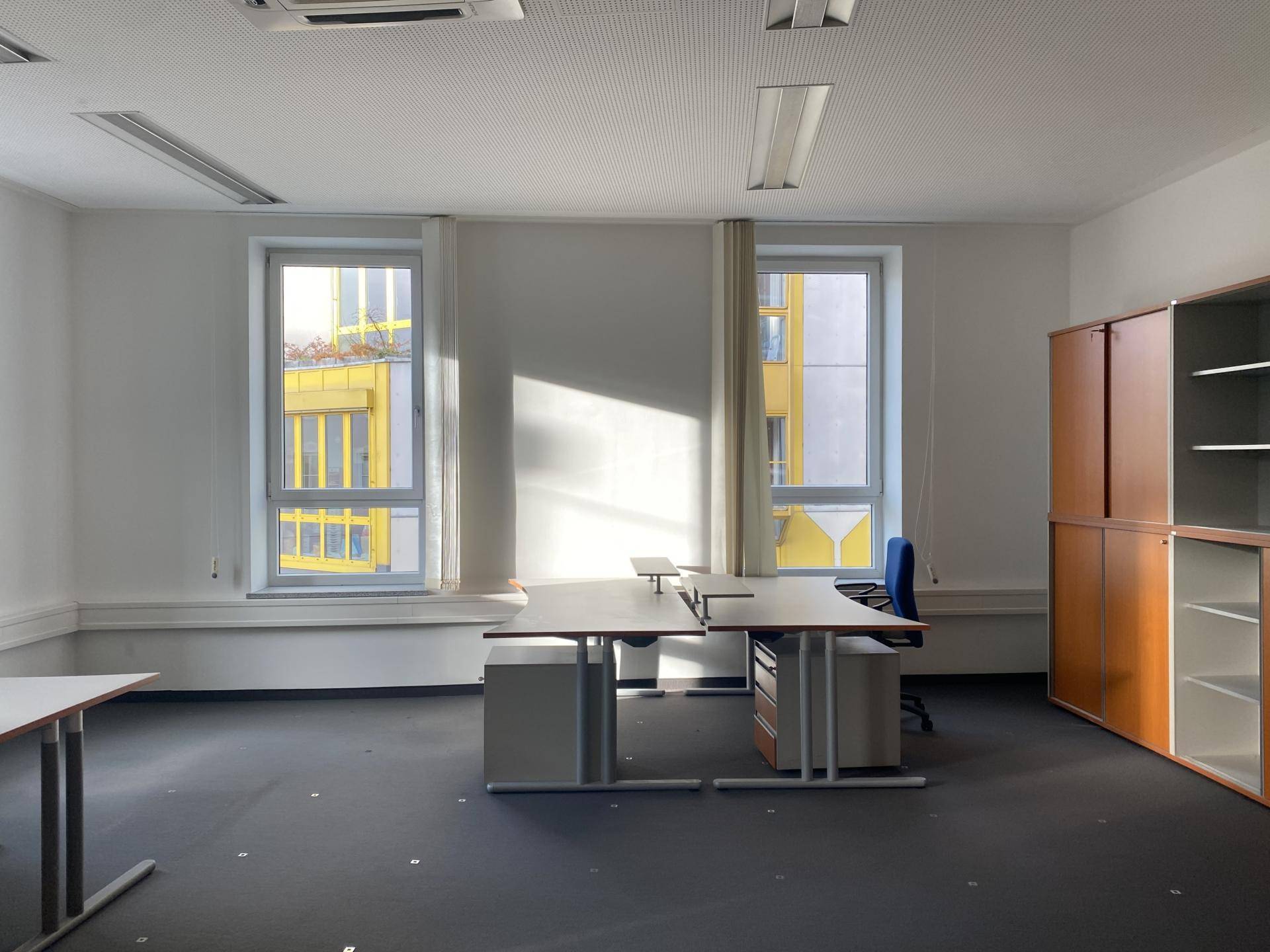 Heller Büroraum mit zwei großen Fenstern, Schreibtischen und Bürostühlen, gut beleuchtet mit Stadtblick.