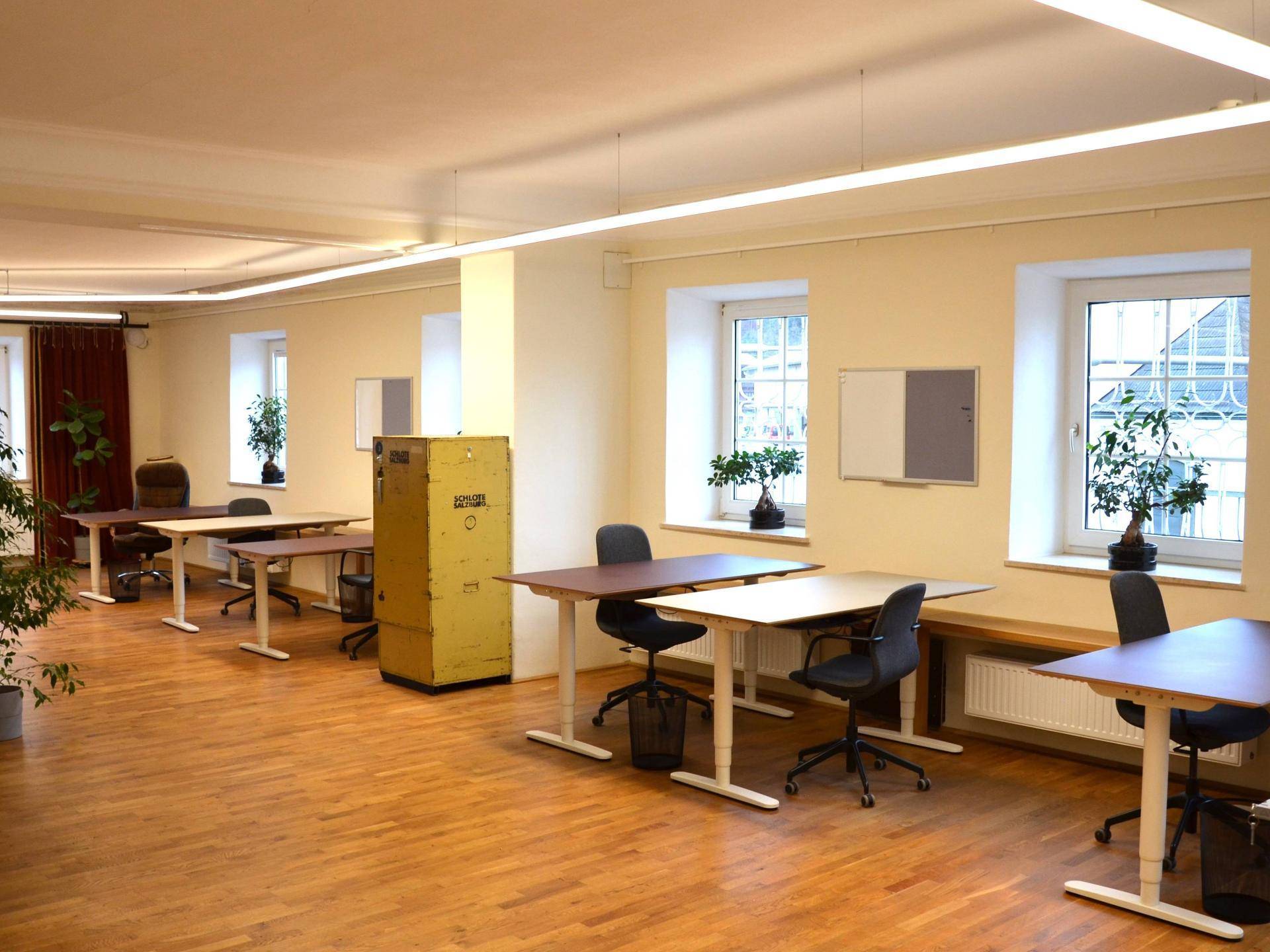 Heller Büroraum mit mehreren Arbeitsplätzen, großen Fenstern und Holzboden, ausgestattet mit Heizkörpern.