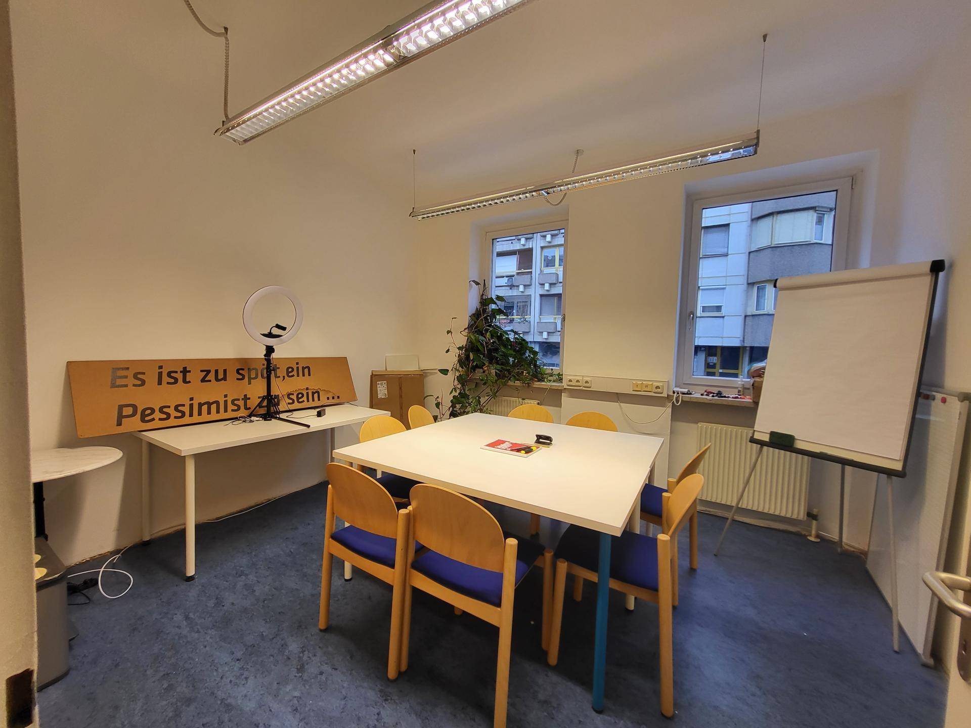 Besprechungsraum mit einem großen Tisch, Stühlen und einem Whiteboard, beleuchtet von Deckenleuchten und Fenstern.