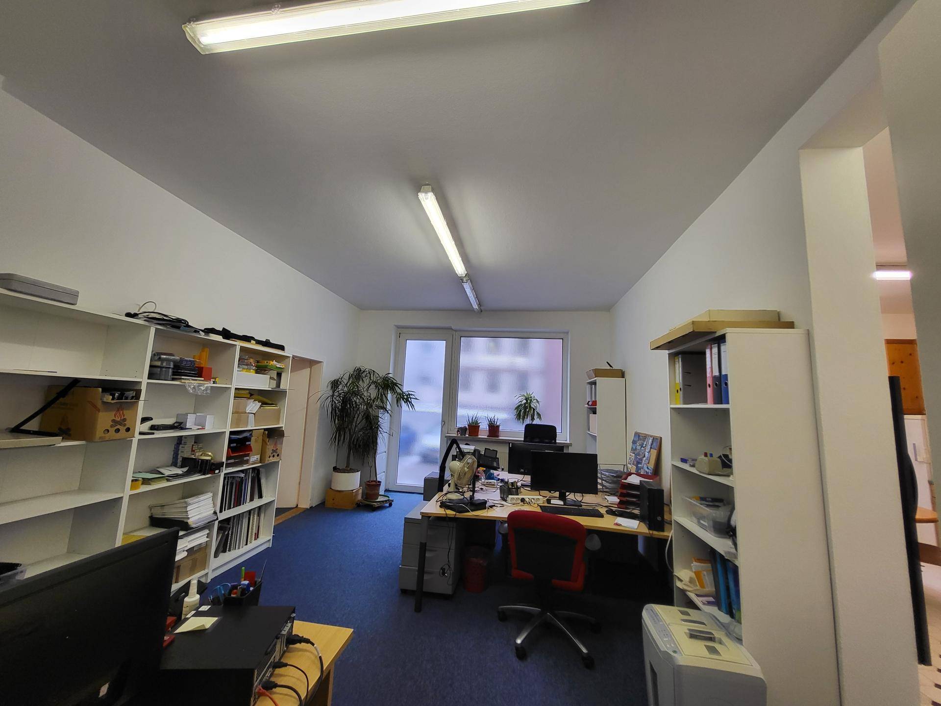 Geräumiges Büro mit mehreren Arbeitsplätzen, Regalen und einem großen Fenster, das viel Tageslicht hereinlässt.