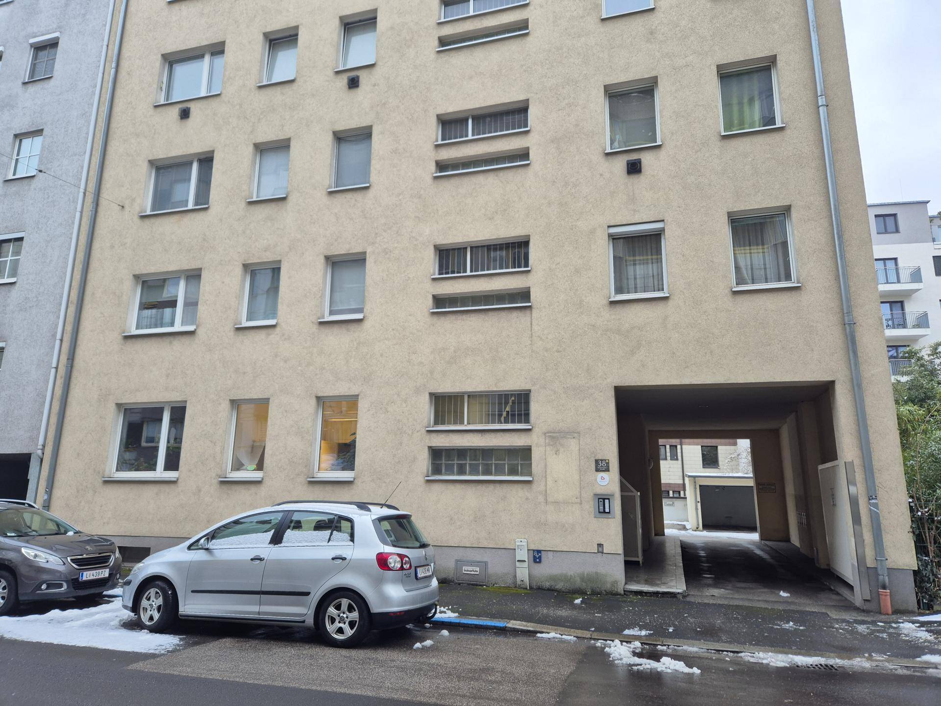 Ansicht eines mehrstöckigen Gebäudes mit beiger Fassade, Fenstern und einem Durchgang zum Innenhof oder Parkplatz.
