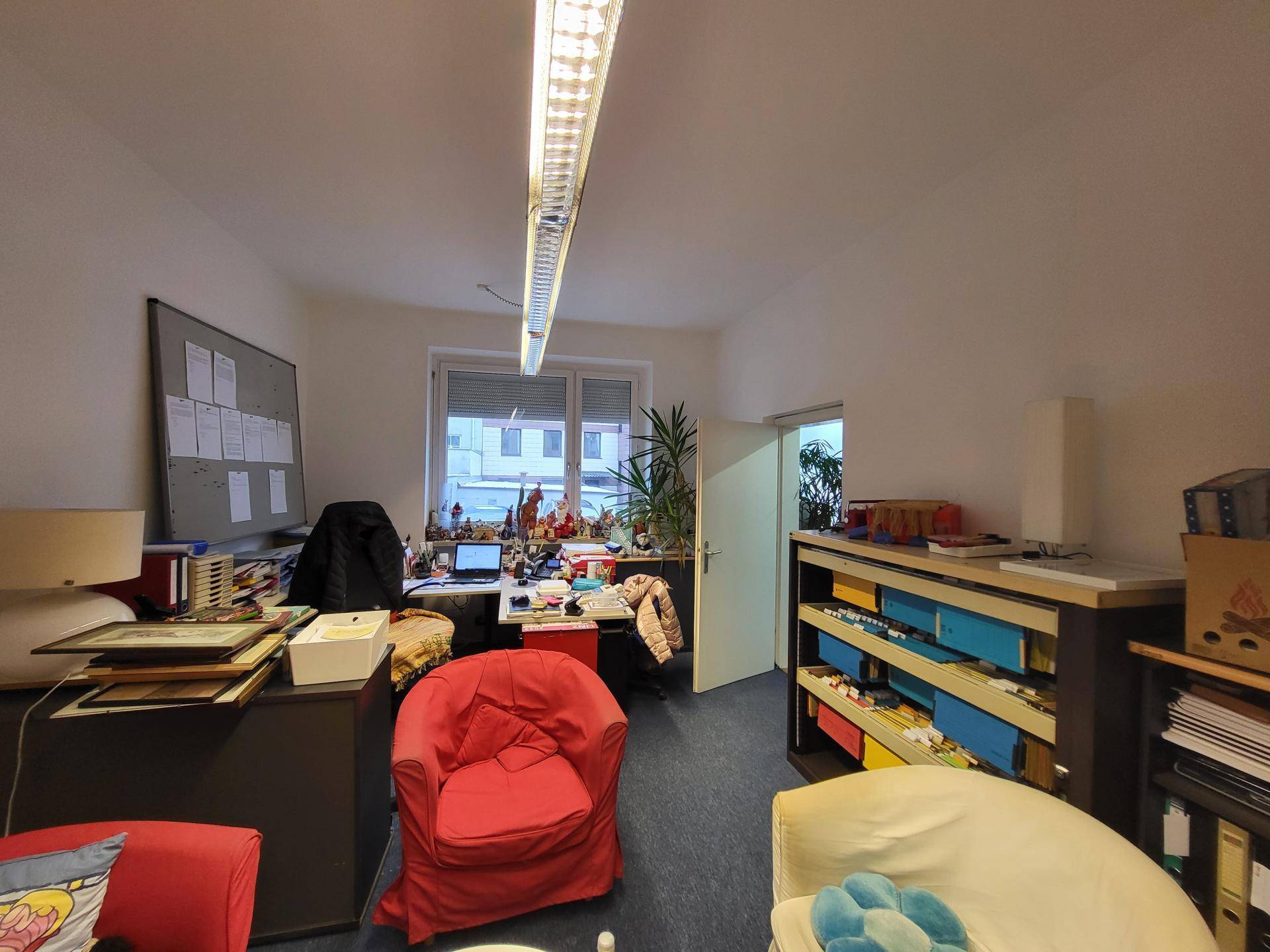 Büroarbeitsplatz mit Schreibtisch, Computer und bunten Ablagen, beleuchtet von einer Deckenleuchte und Tageslicht.