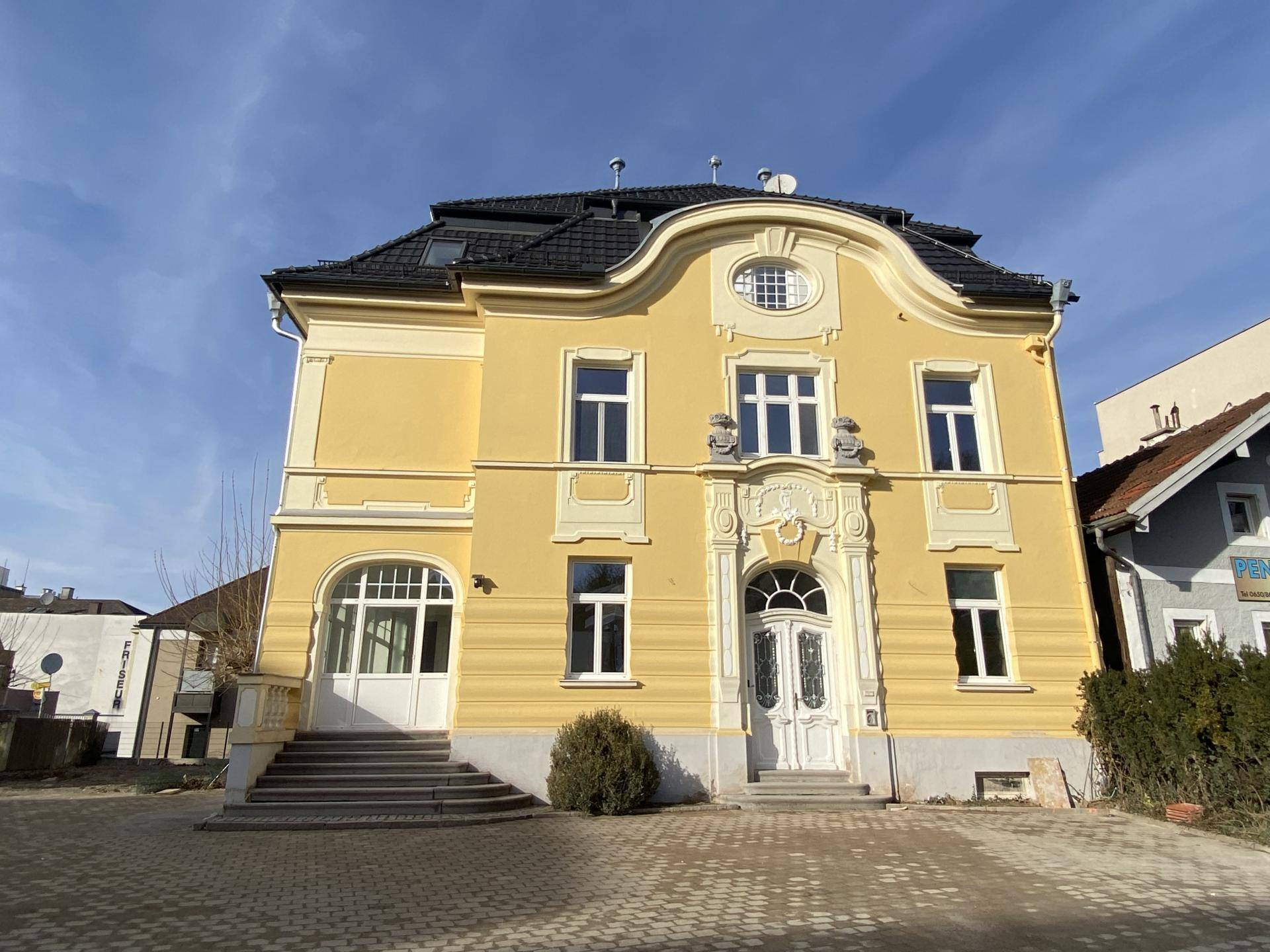 Historisches, gelb gestrichenes Gebäude mit aufwendiger Fassadenverzierung, großen Fenstern und einem gepflasterten Vorplatz.