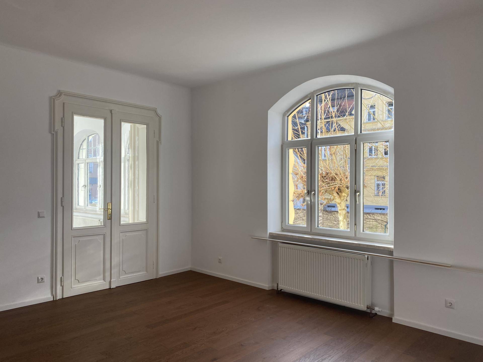 Geräumiger, heller Raum mit Holzboden, hohen Fenstern und klassischer Flügeltür.