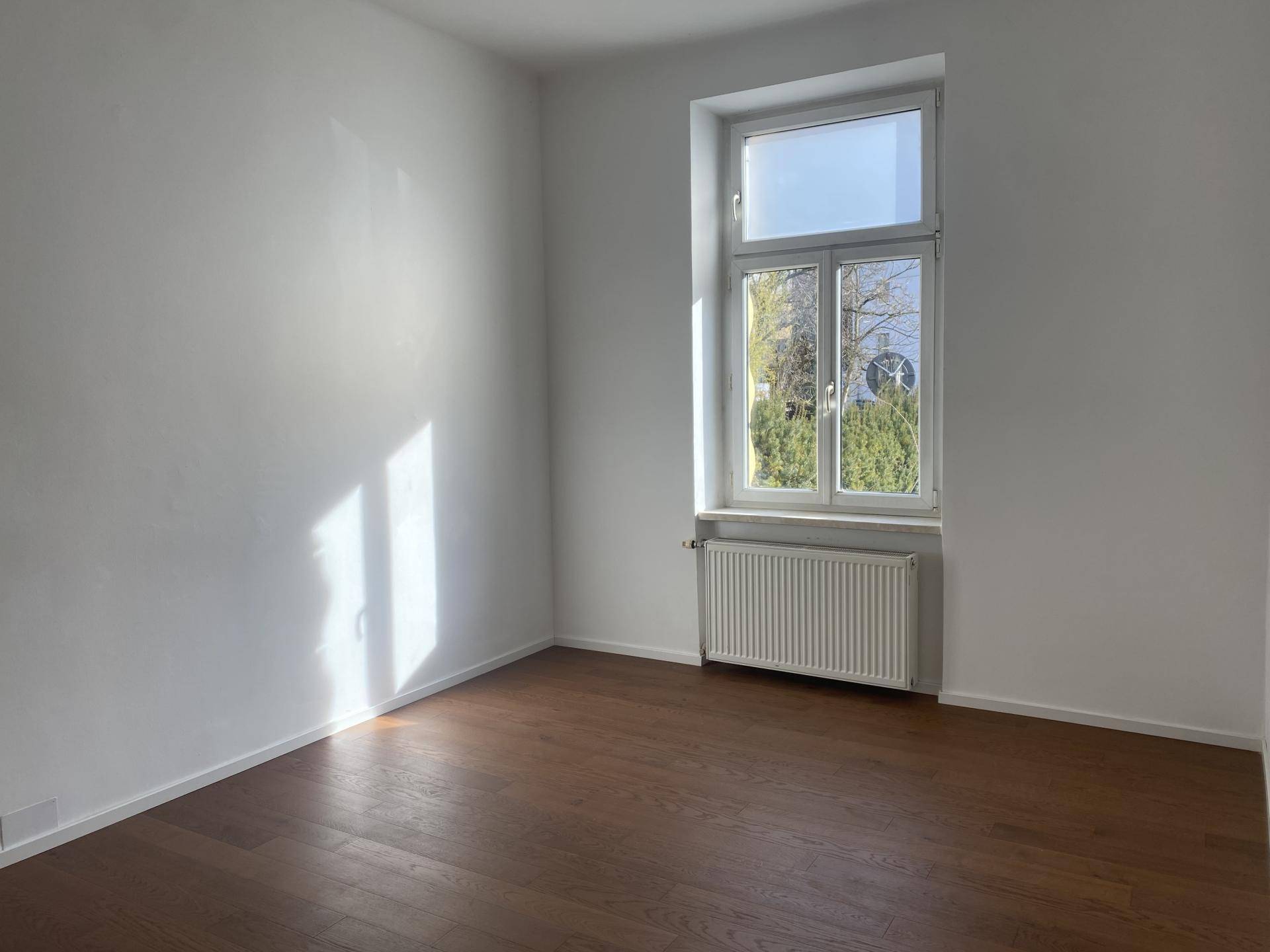 Schlichter Raum mit Holzboden, einem großen Fenster und einem Heizkörper, der viel Tageslicht bietet.