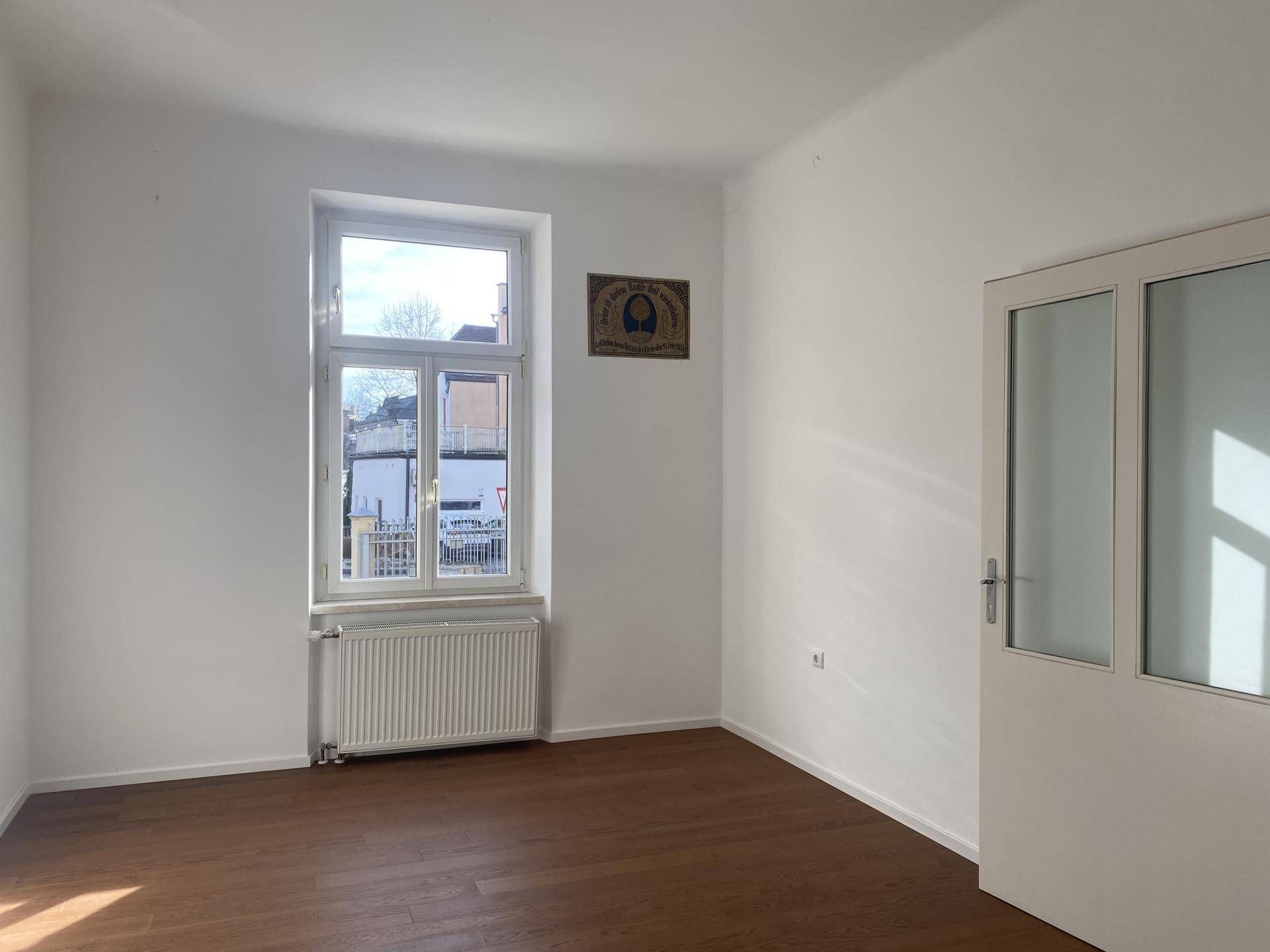 Leerer Raum mit Holzboden, einem Fenster und einer Glastür, die den Zugang zu einem weiteren Bereich ermöglicht.