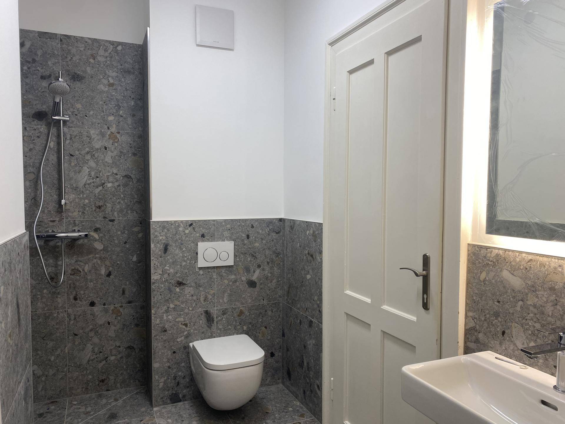 Modernes Badezimmer mit grauen Fliesen, einer ebenerdigen Dusche, wandhängendem WC und einem Waschbecken mit beleuchtetem Spiegel.