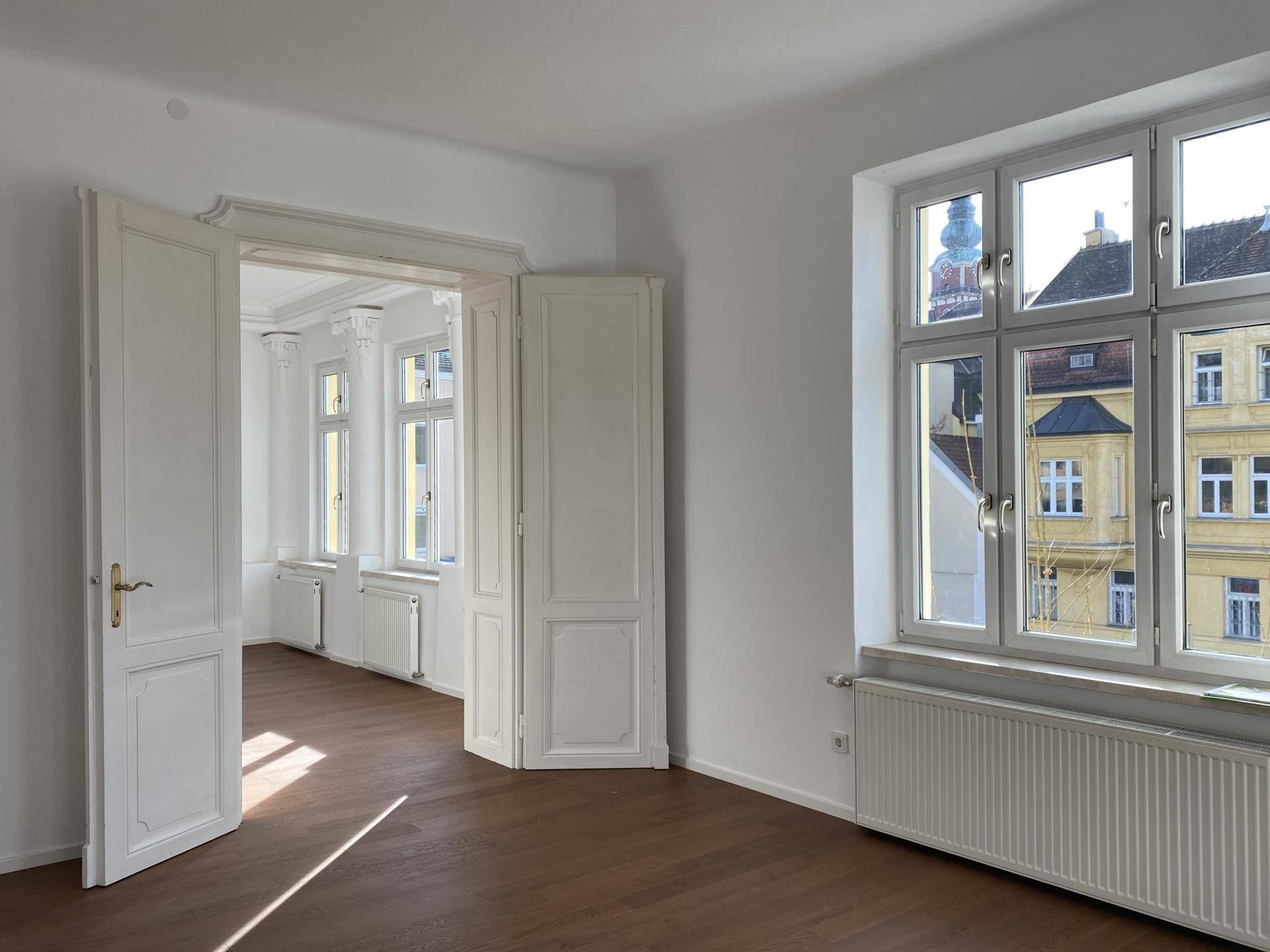 Heller Wohnraum mit Holzboden, großen Fenstern und Blick auf die umliegenden Gebäude.