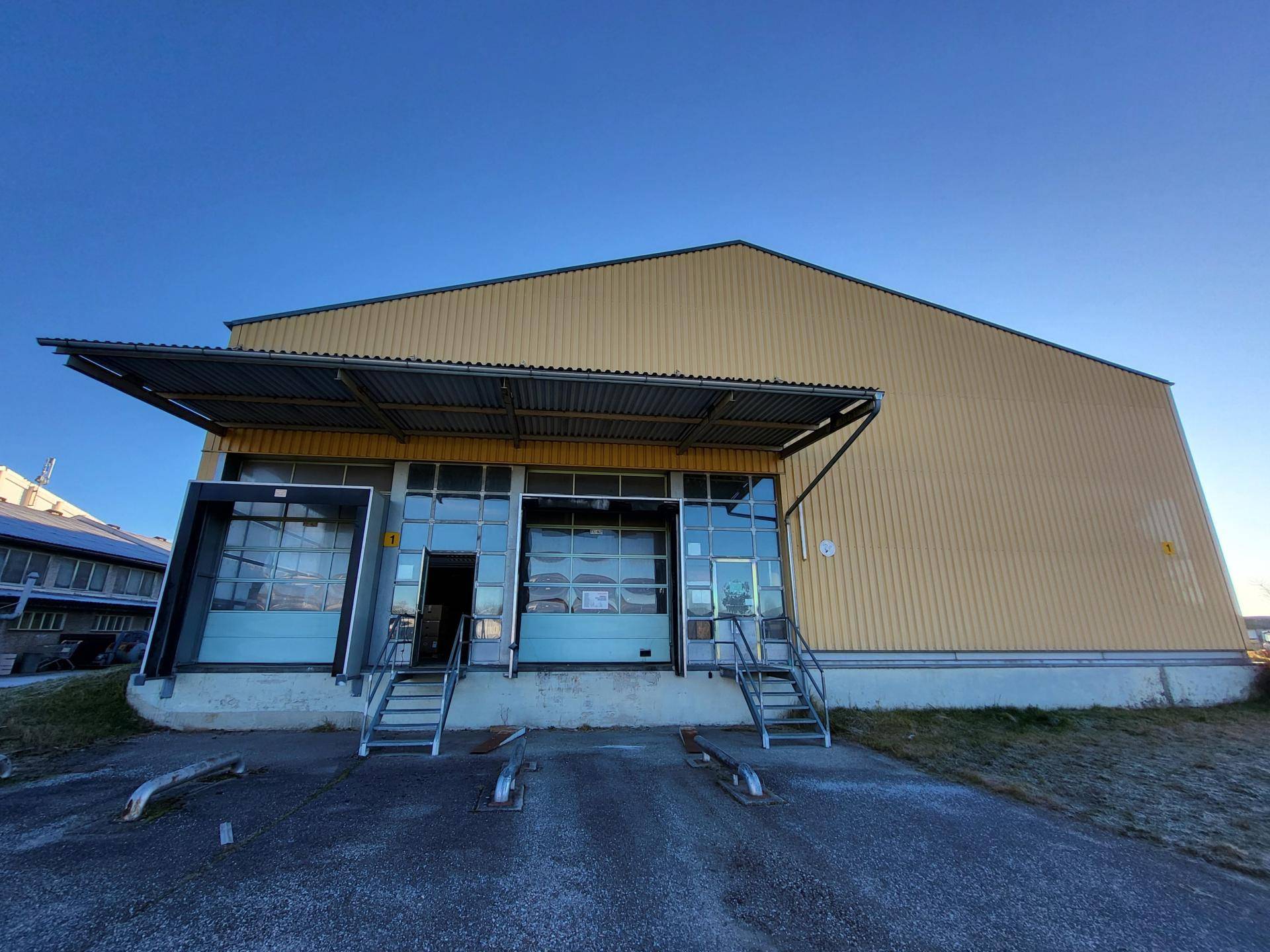 Seitenansicht einer modernen Lagerhalle mit großen Rolltoren und Zugangstreppen unter klarem Himmel.